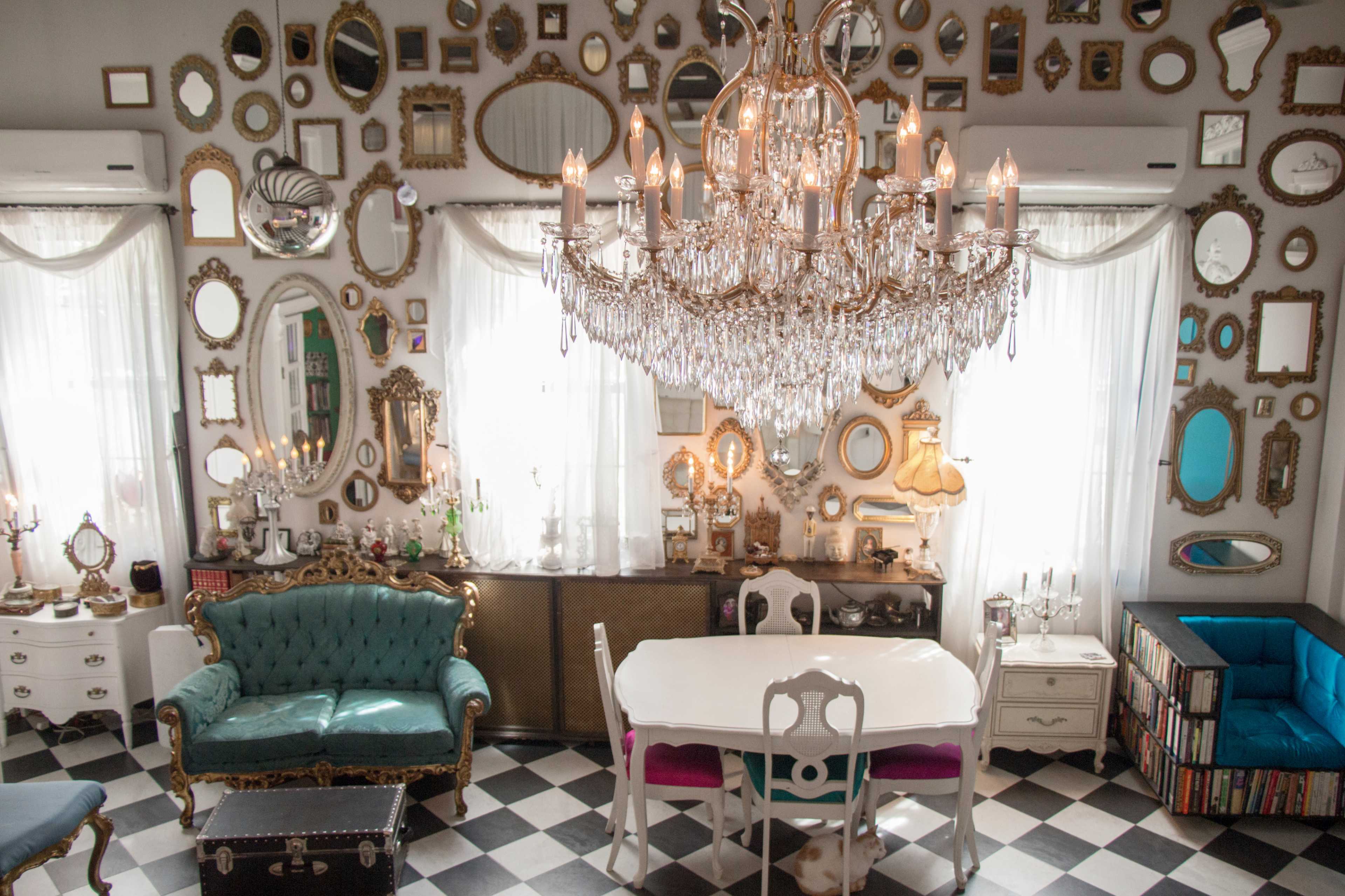 A bright room features a white dining table surrounded by chairs, with a large chandelier hanging above and walls adorned with various mirrors.