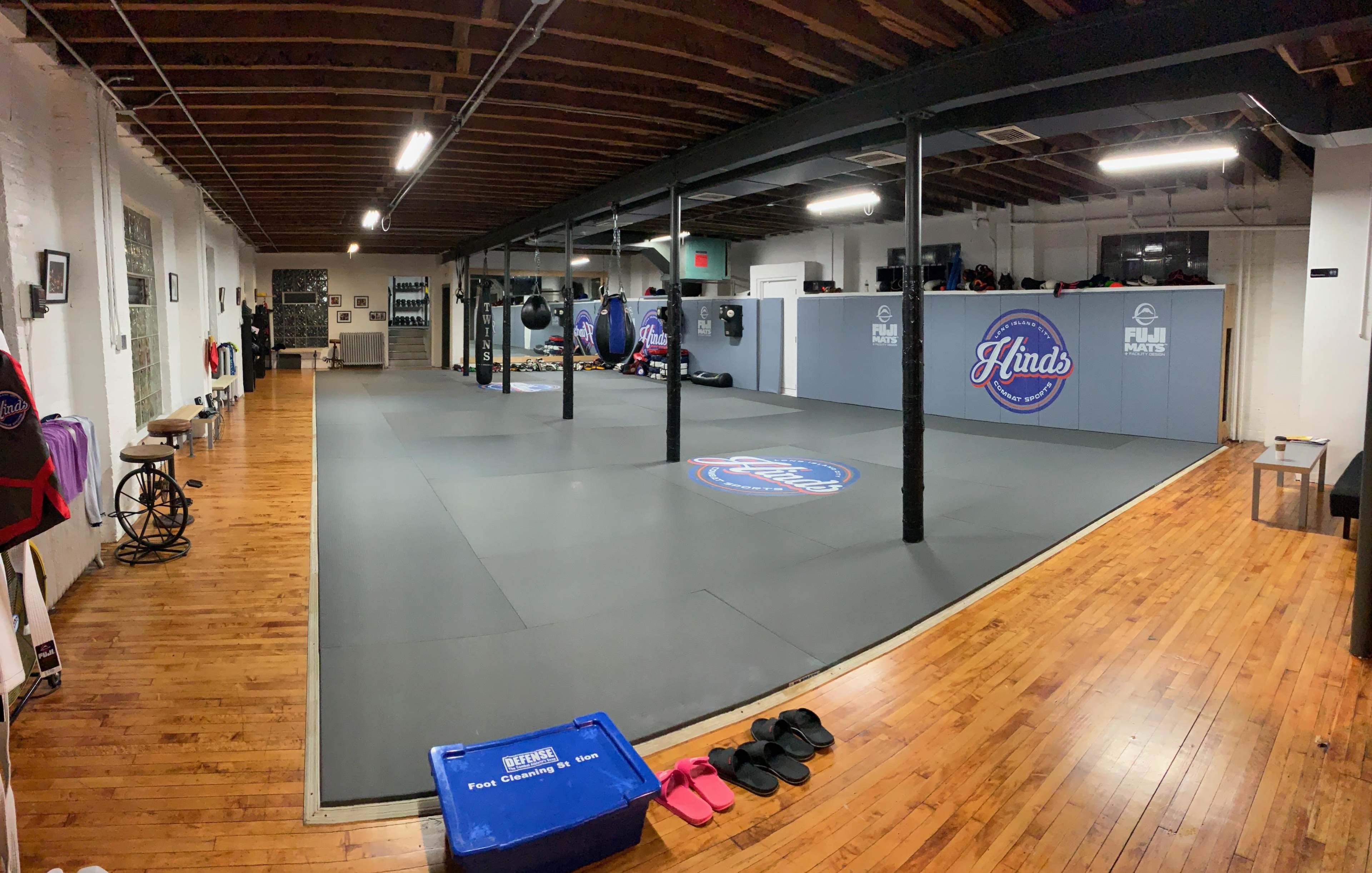 A spacious martial arts studio with padded flooring, hanging punching bags, and a centralized logo on the mat.