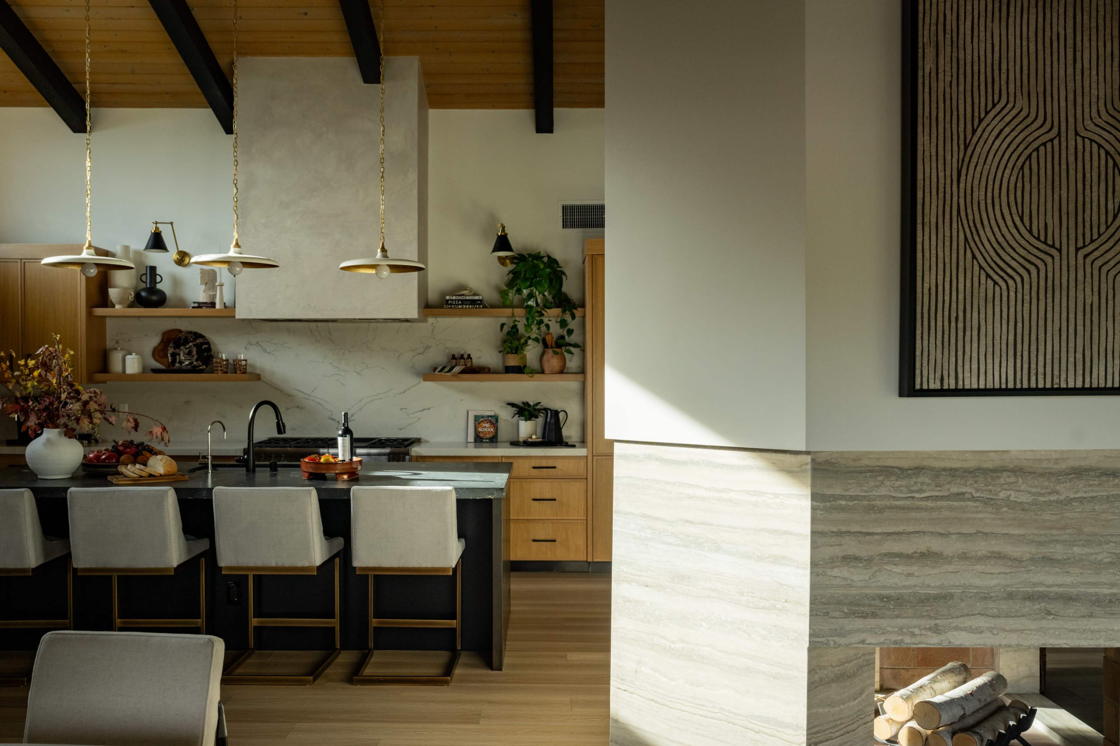 A modern kitchen with wooden beams features a large island with seating, an elegant backsplash, and a fireplace in the adjacent living area.