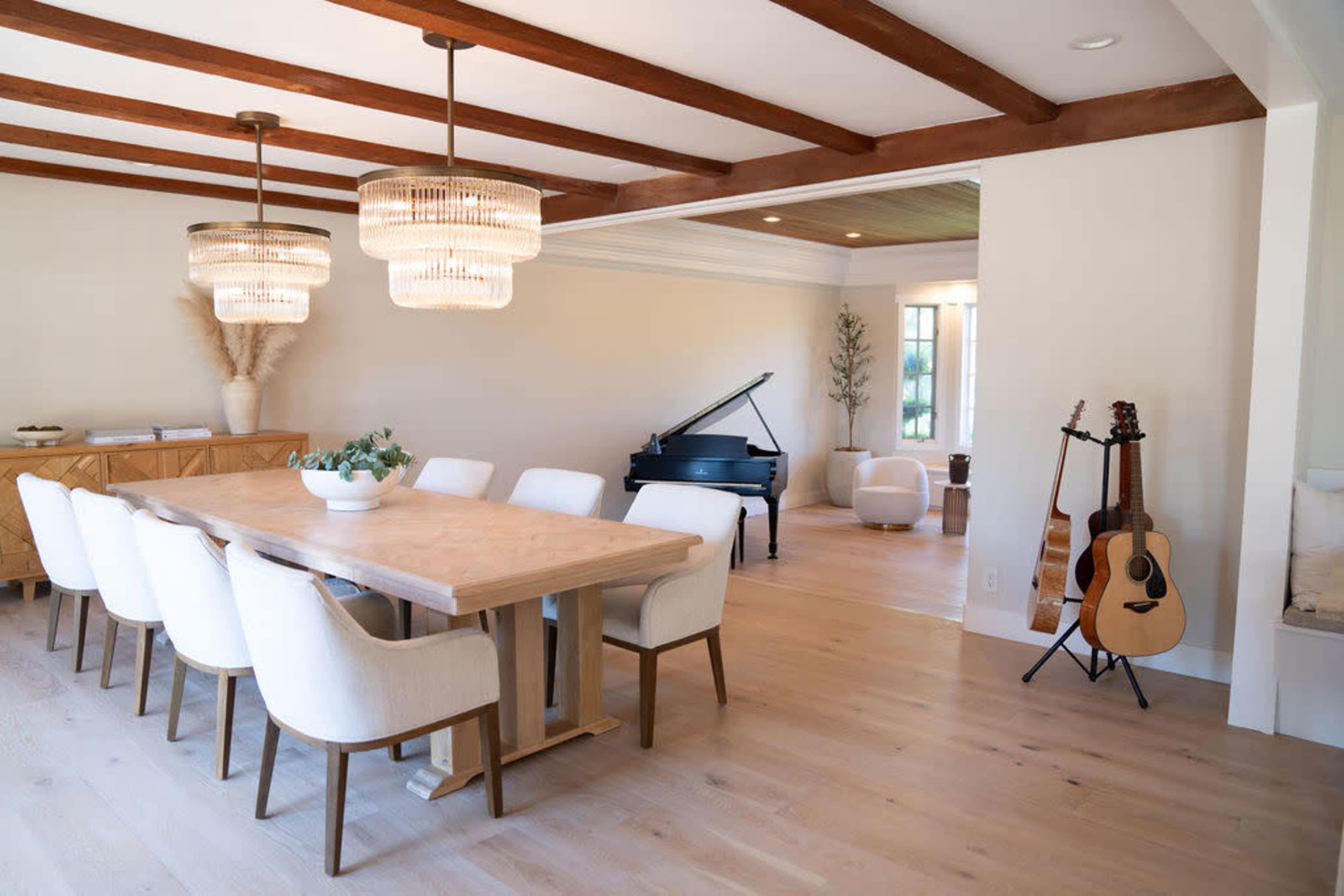 A modern dining area with a large wooden table surrounded by white chairs, a grand piano in the corner, and warm pendant lights overhead.