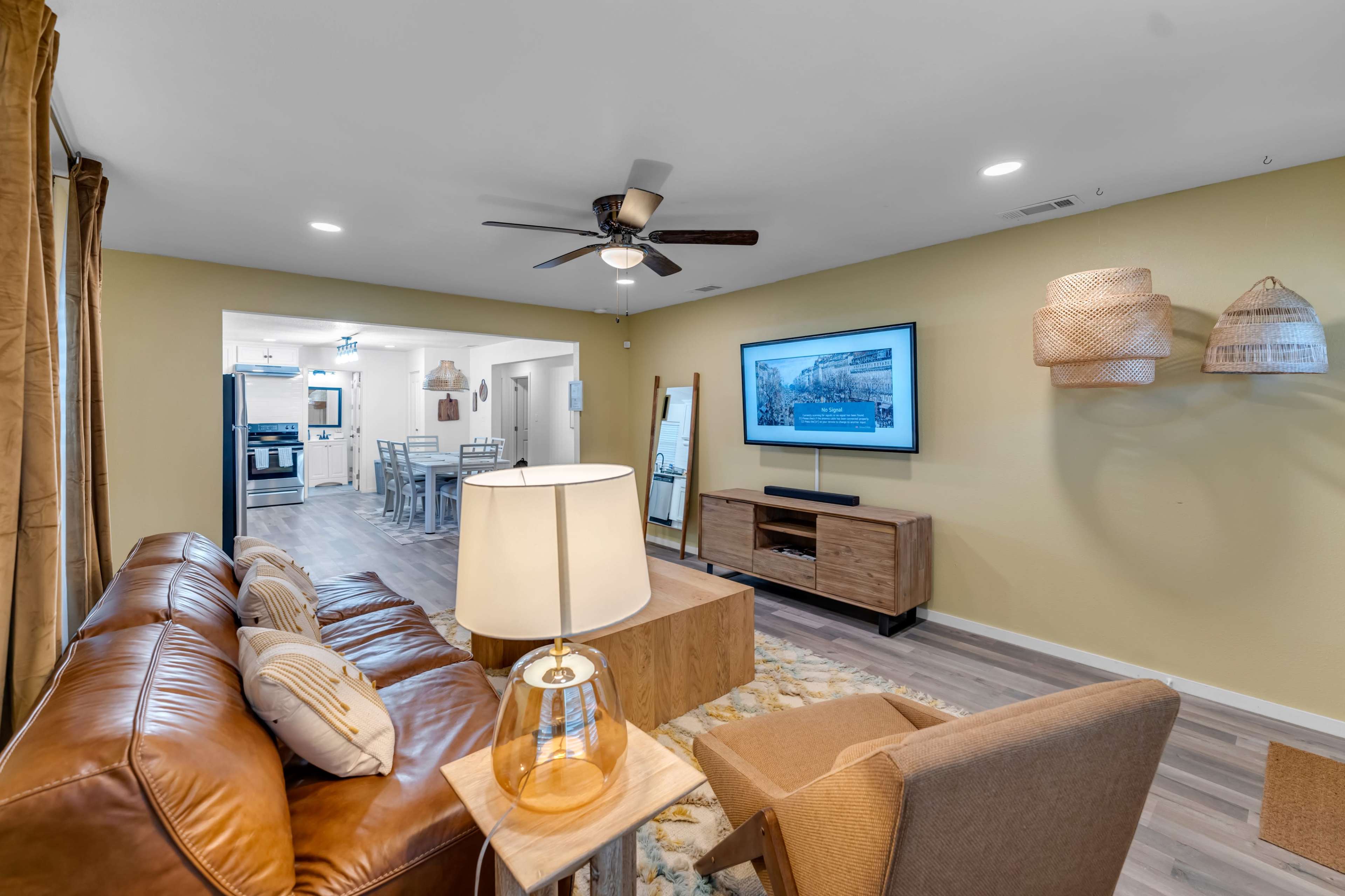 The image shows a living room with a brown leather couch, a wooden coffee table, a ceiling fan, a television mounted on the wall, and light-colored walls and flooring.