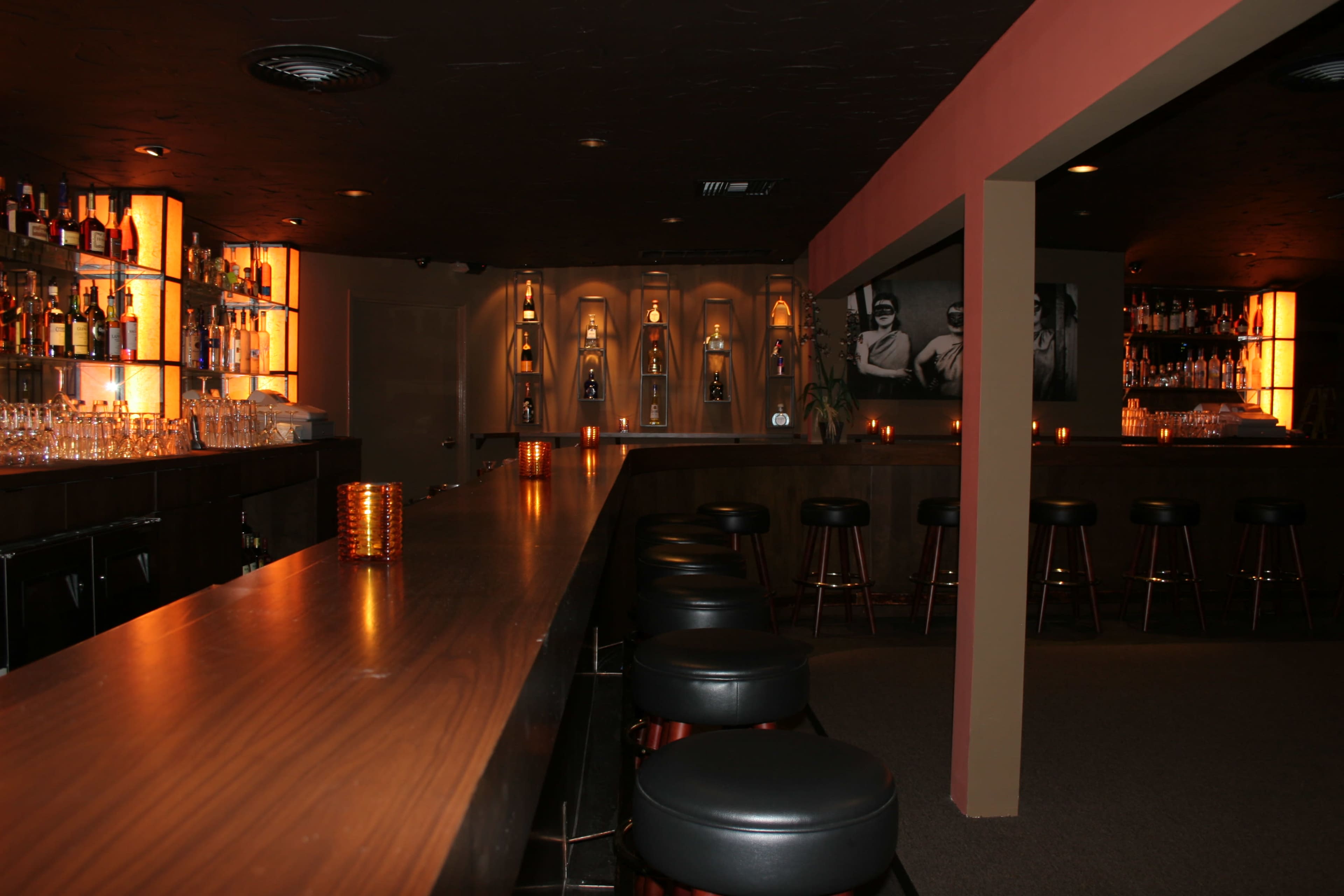 A dimly lit bar features a long wooden counter lined with black stools and illuminated shelves displaying various bottles.