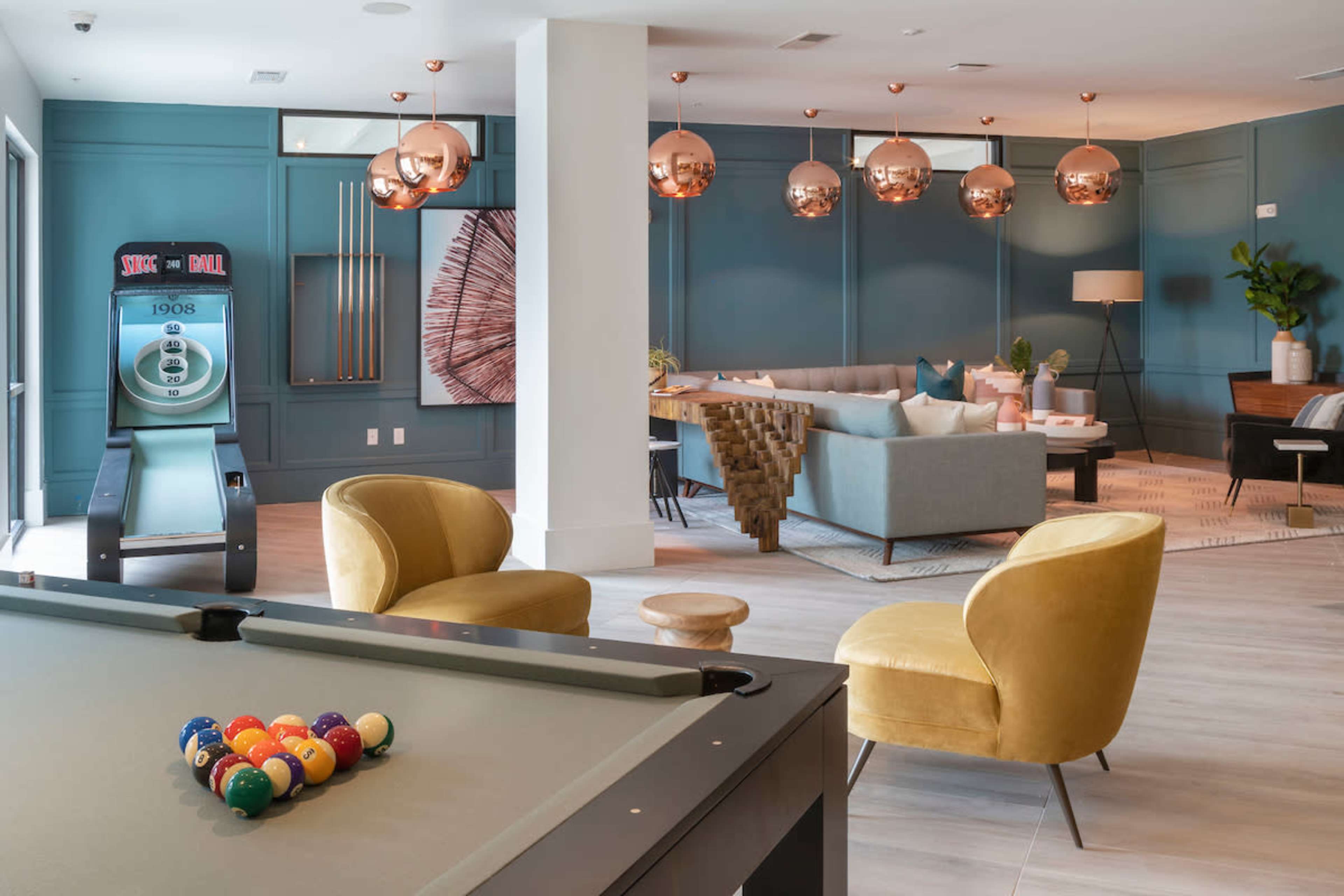 A modern lounge area features a billiard table, two yellow accent chairs, a gray sofa, and decorative pendant lights hanging from the ceiling.