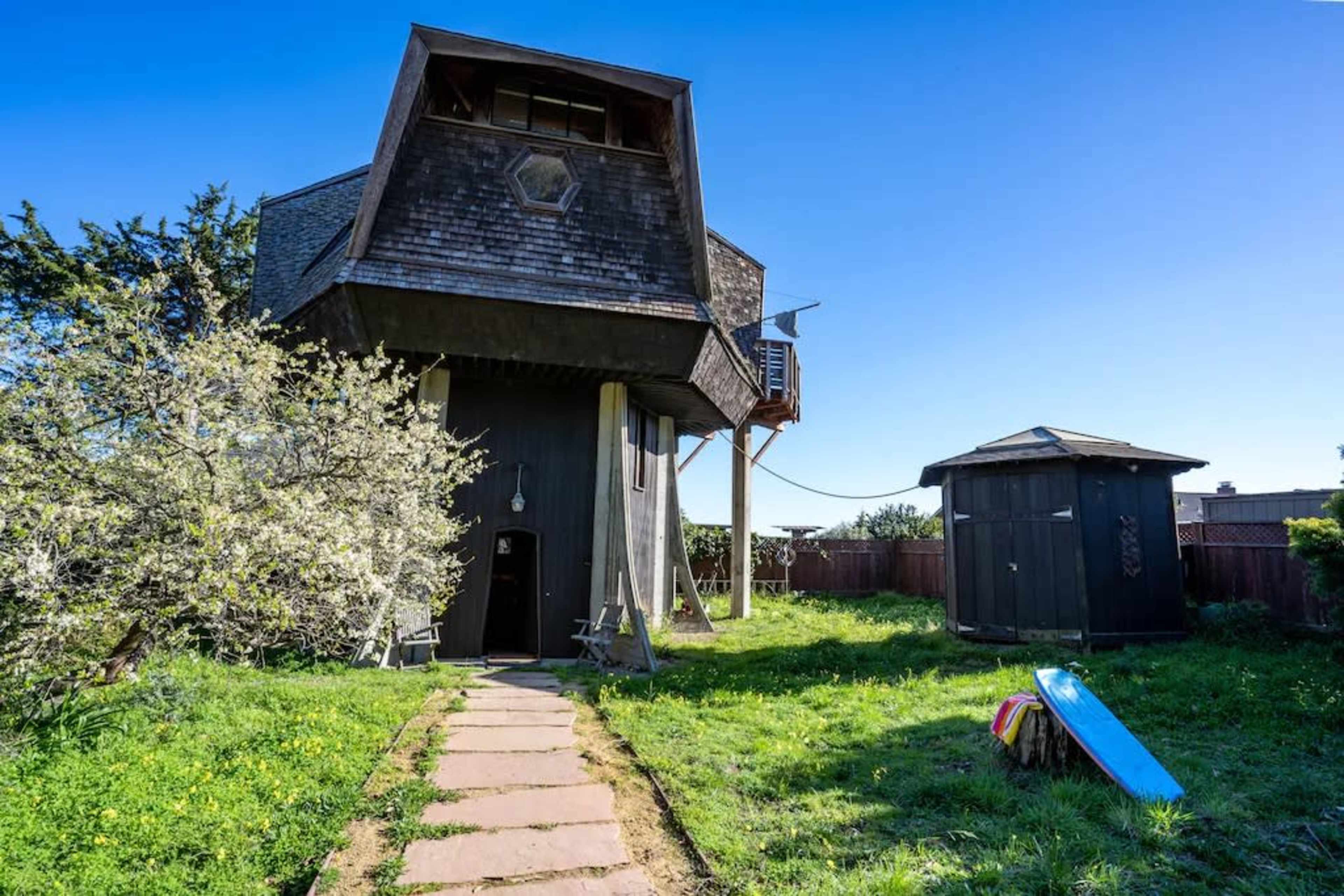 A unique, multi-level house with an angular roof stands in a grassy yard, accompanied by a small shed and a surfboard leaning against a bench.