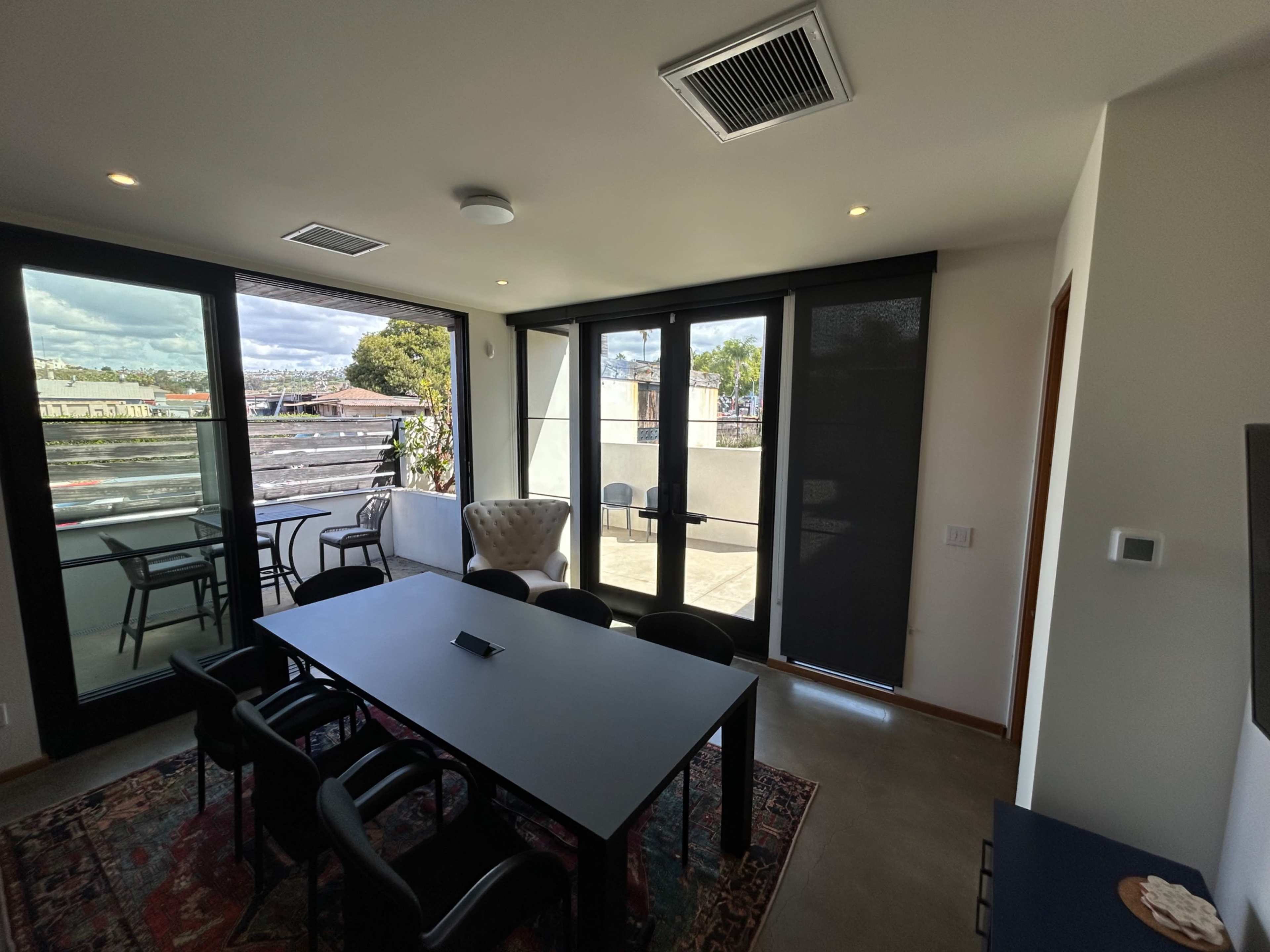 The image shows a modern indoor space with a dining table and chairs, large windows leading to a balcony, and a comfortable armchair.