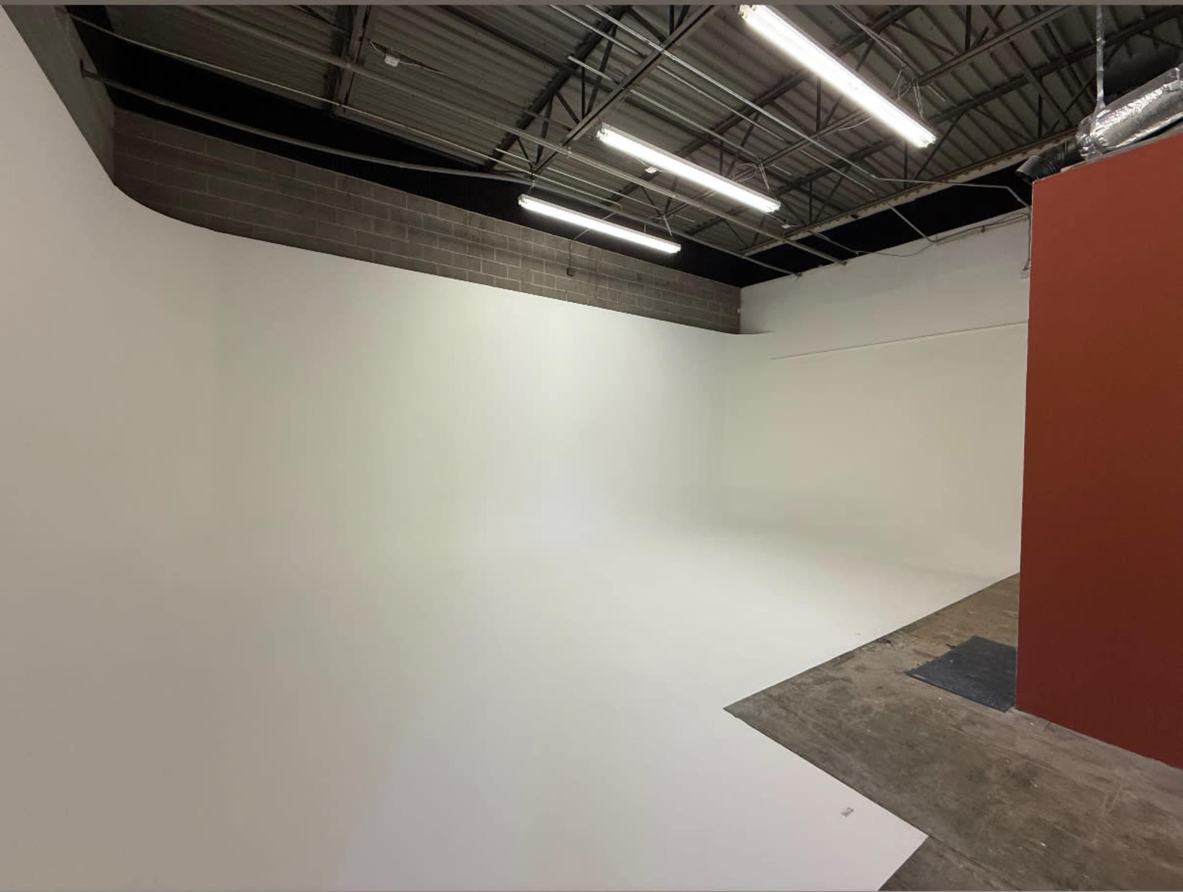 The image shows a spacious, empty studio with white walls and floor, featuring exposed ceilings with fluorescent lights.