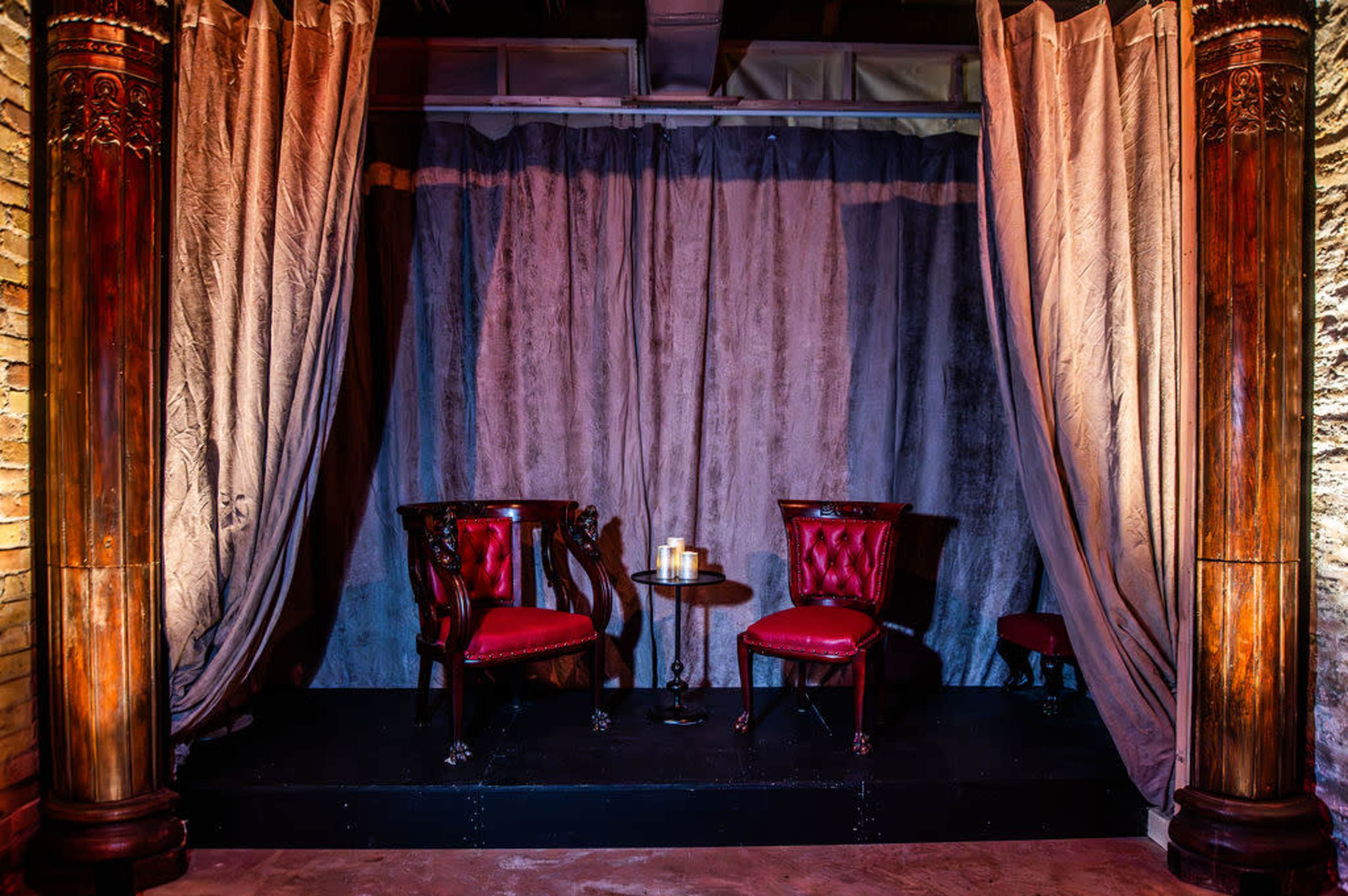 The image shows a stage with two red upholstered chairs flanking a small round table, set against a backdrop of gray curtains and wooden columns.