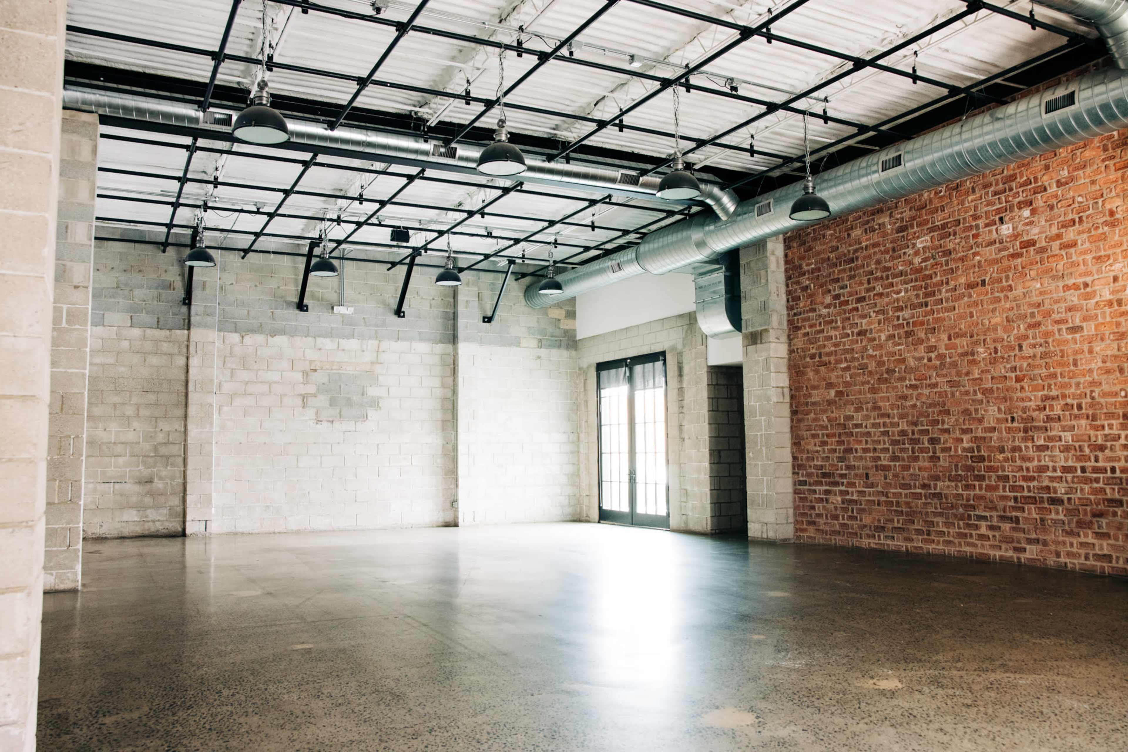 The image shows an empty industrial-style interior space featuring concrete and exposed brick walls, large windows, and a high ceiling with visible metal beams.