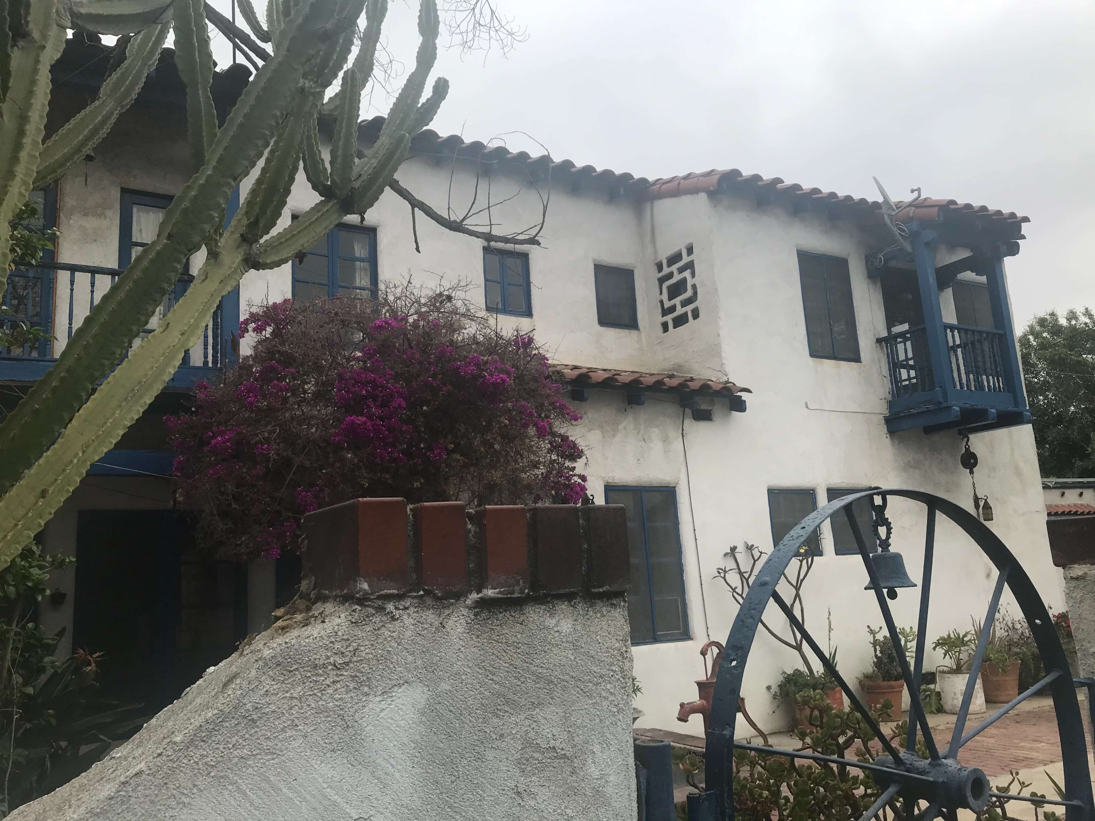 A two-story building with blue railings and a stucco exterior is surrounded by a cactus and flowering plants.