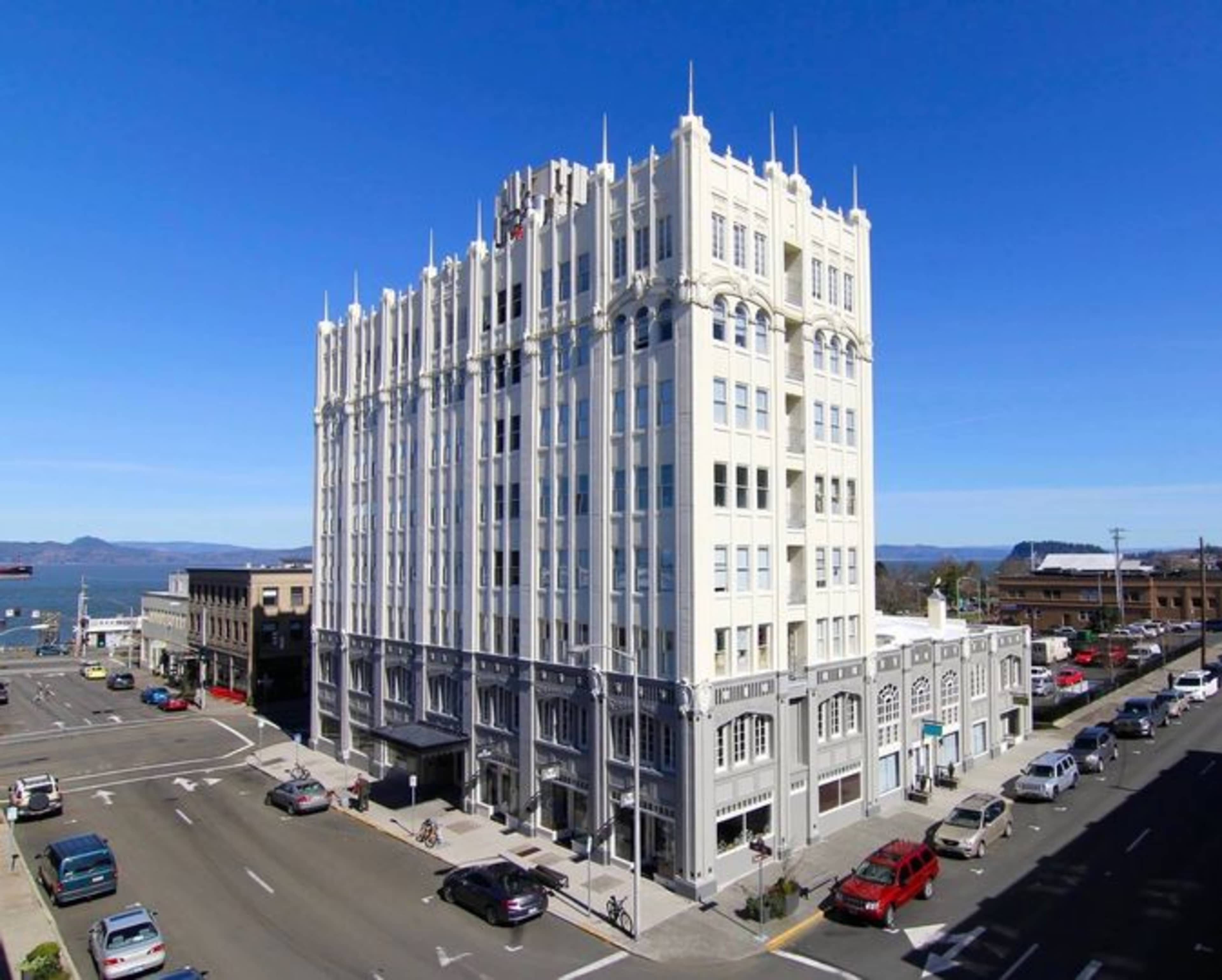 A tall, historic white building with ornate architectural details stands at the intersection of two streets near a body of water.