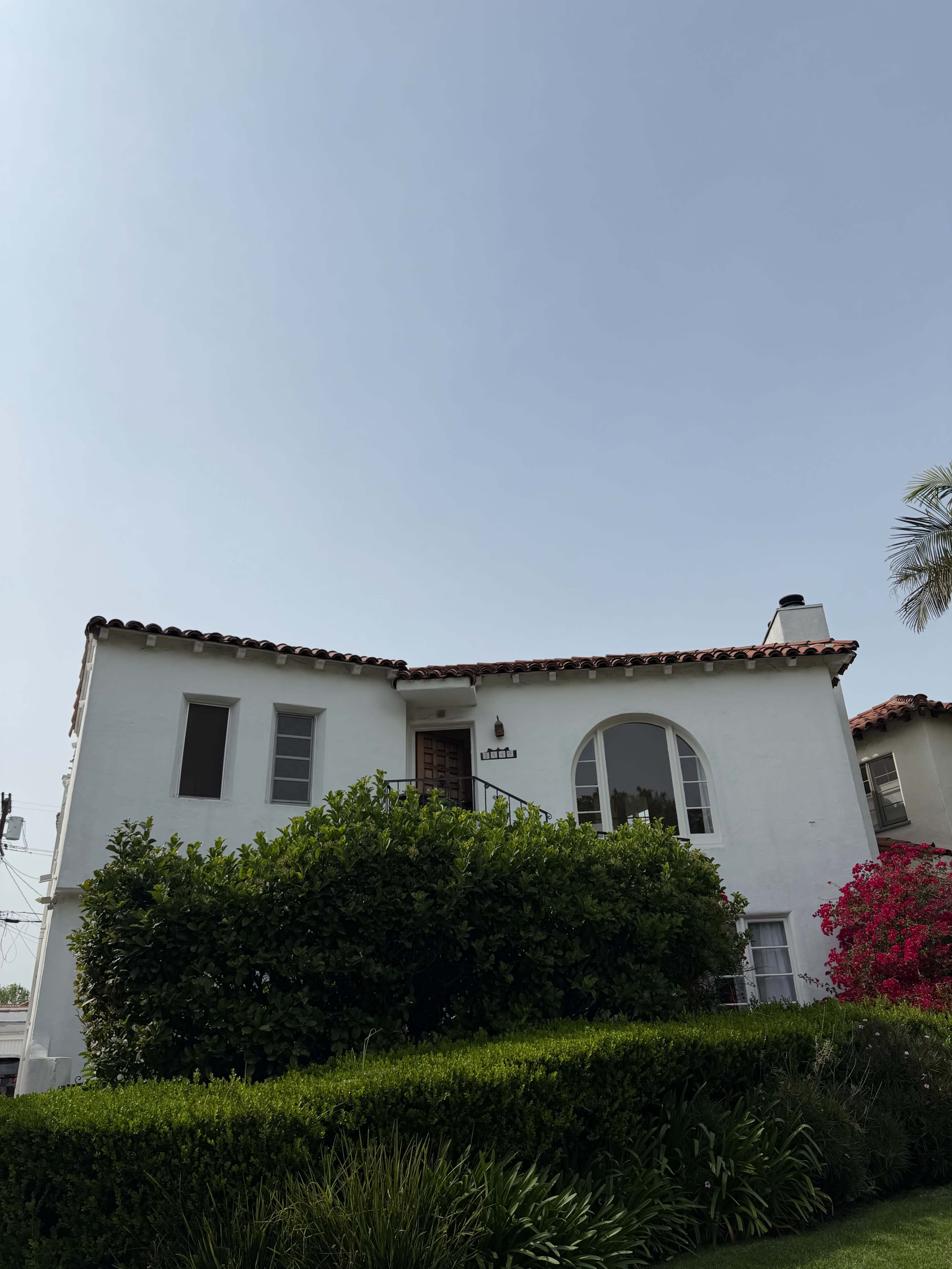 A two-story white house with a red-tiled roof is surrounded by green shrubs and a blooming pink plant under a clear sky.