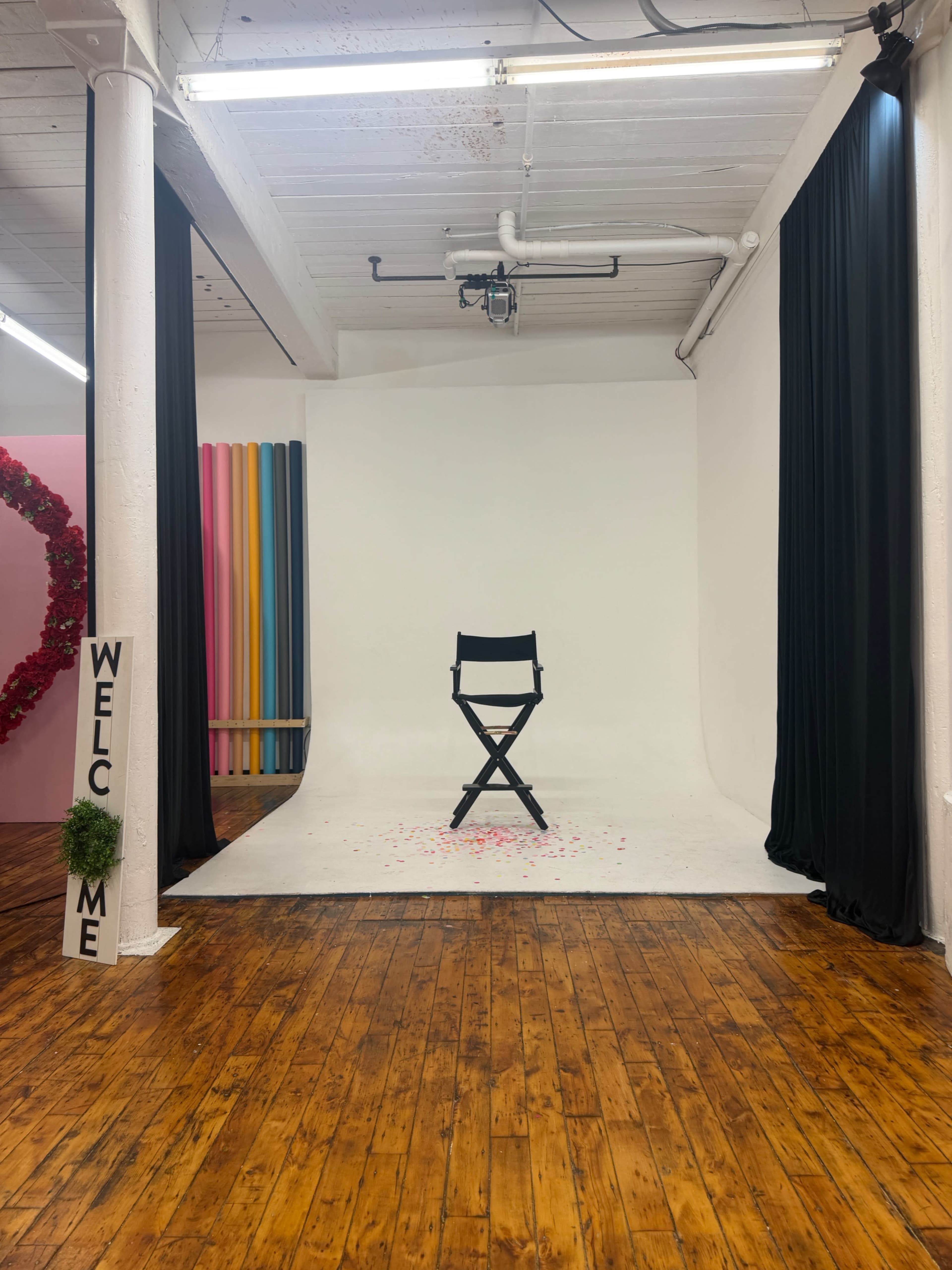 The image features a studio setting with a black director's chair on a white backdrop, surrounded by colorful vertical strips and a decorative sign that says "WELCOME ME."