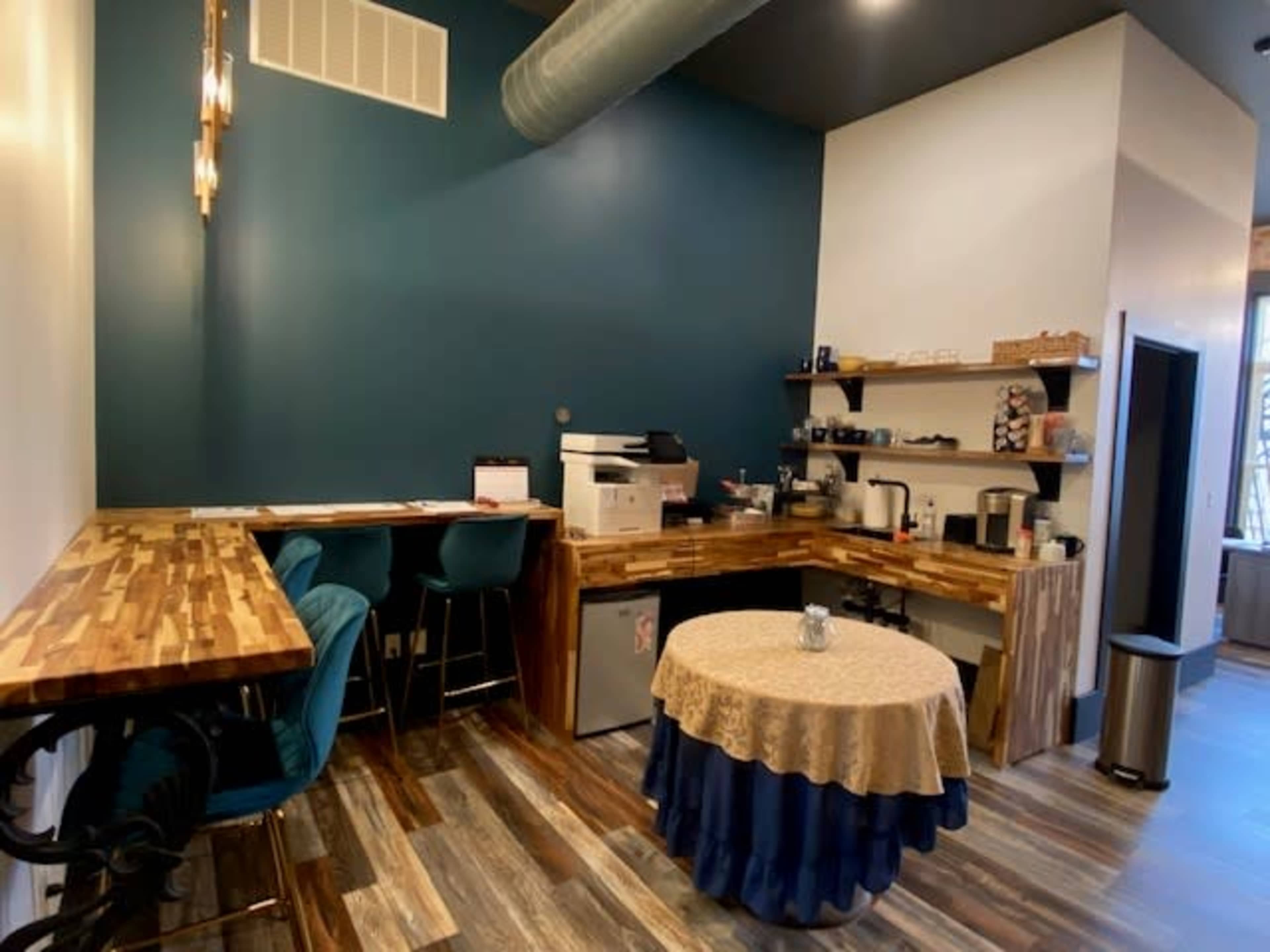 A small café space with a wooden table, teal walls, a countertop with various coffee equipment, and a round table covered with a lace tablecloth.