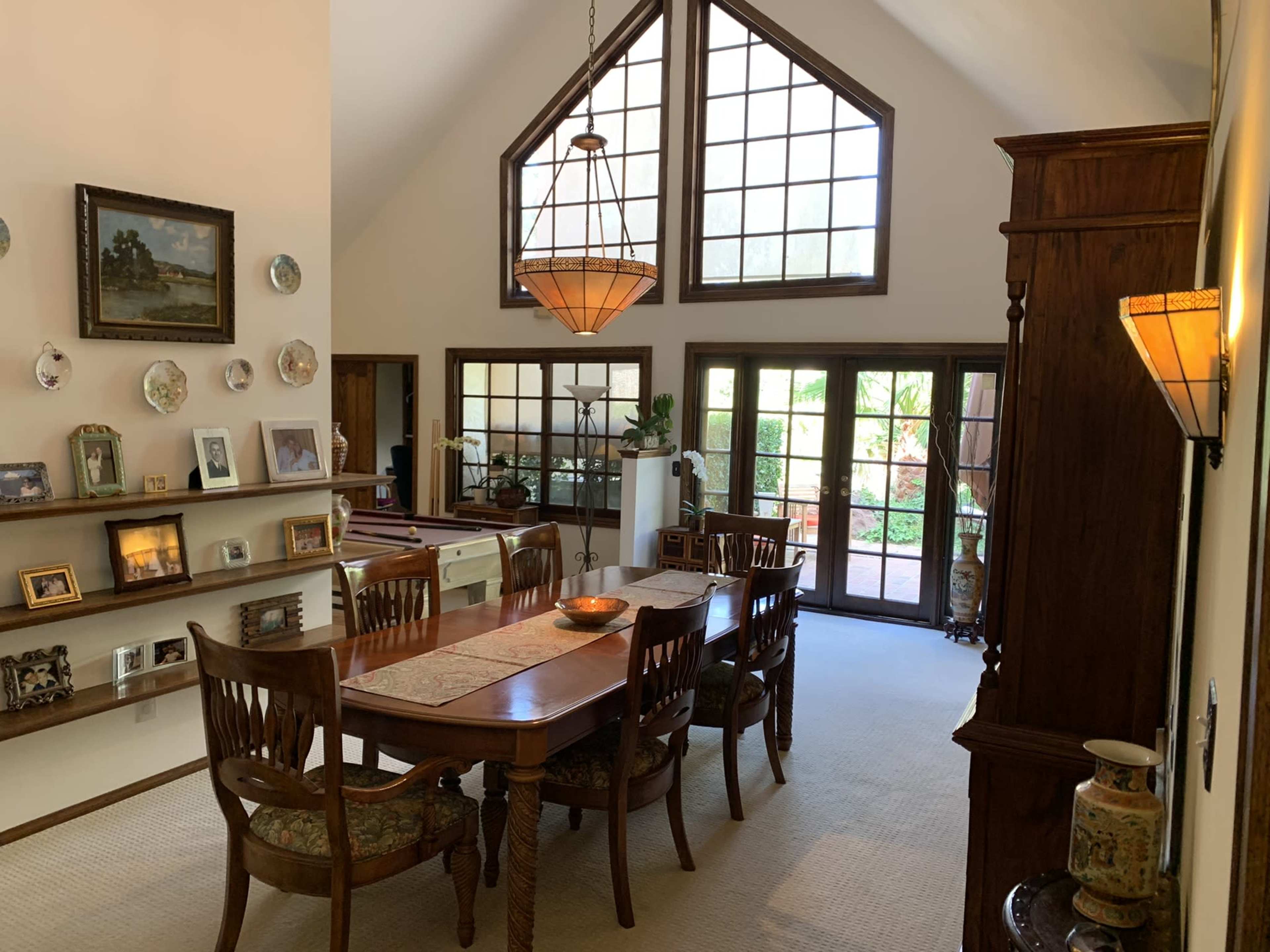 A spacious dining area features a large wooden table surrounded by chairs, with a high ceiling and multiple windows allowing natural light to enter.