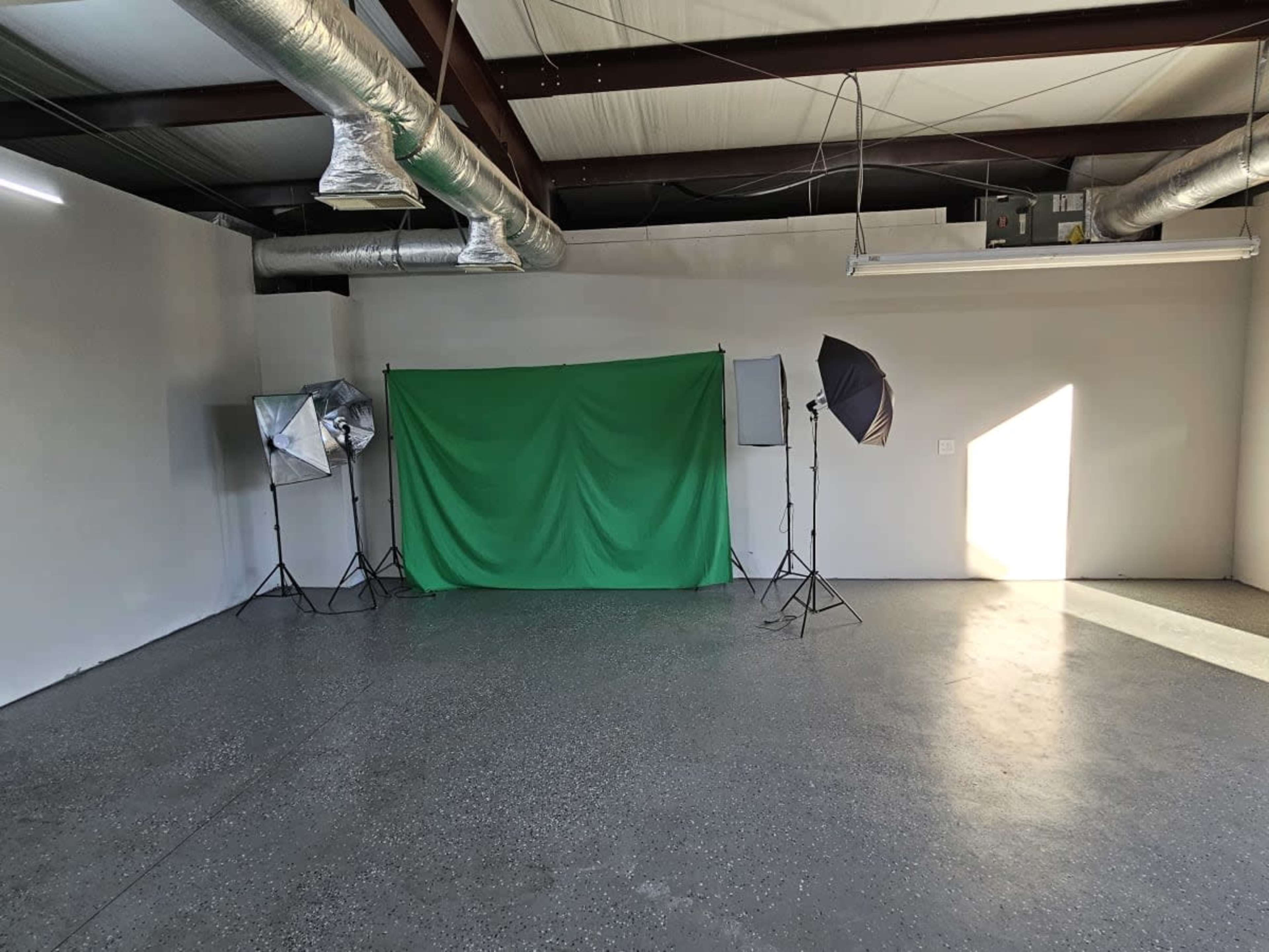 An empty studio space with a green backdrop, two softbox lights, and a window letting in natural light.