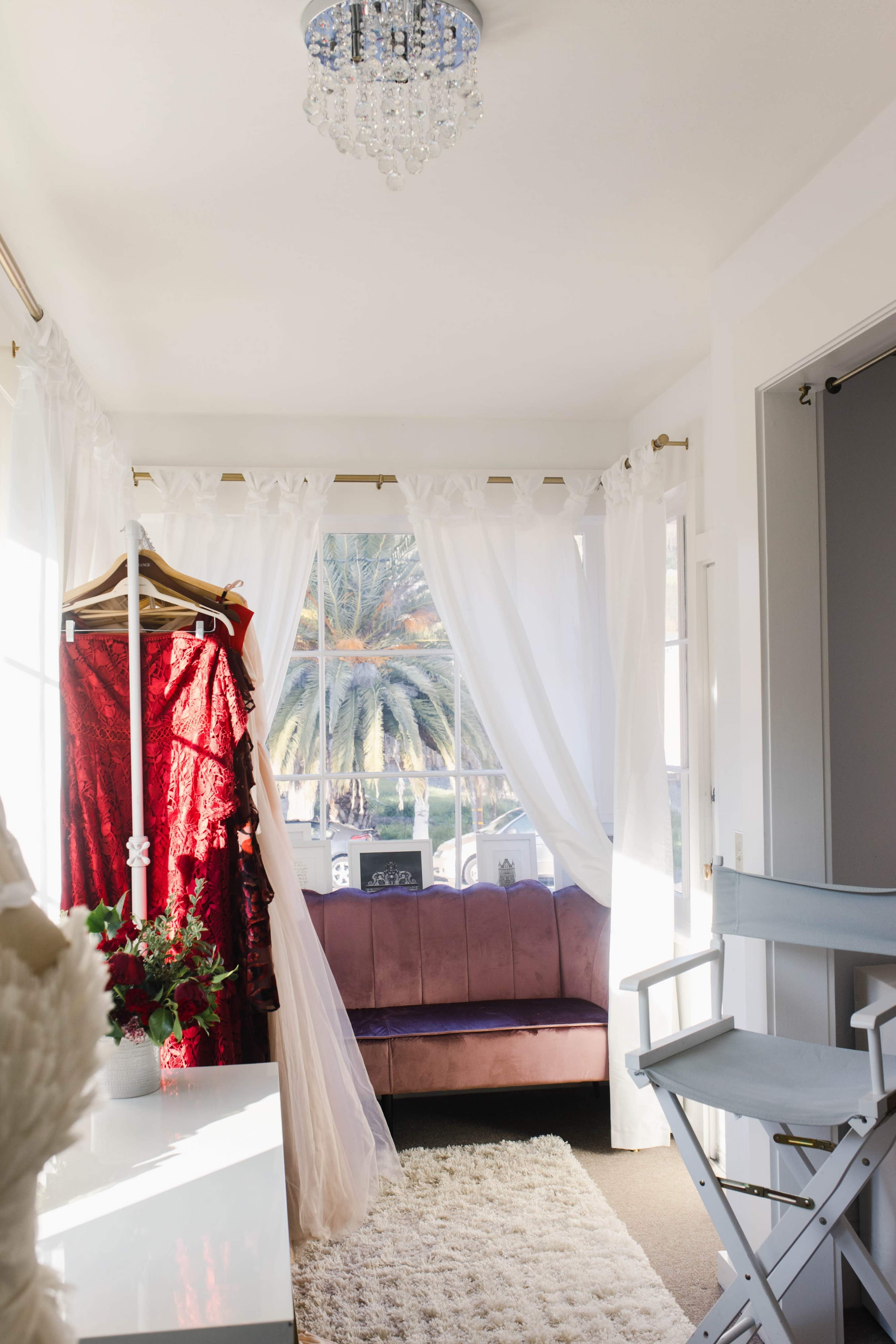 A well-lit room features a plush couch and a rack displaying elegant dresses, with sheer curtains framing a view of palm trees outside.