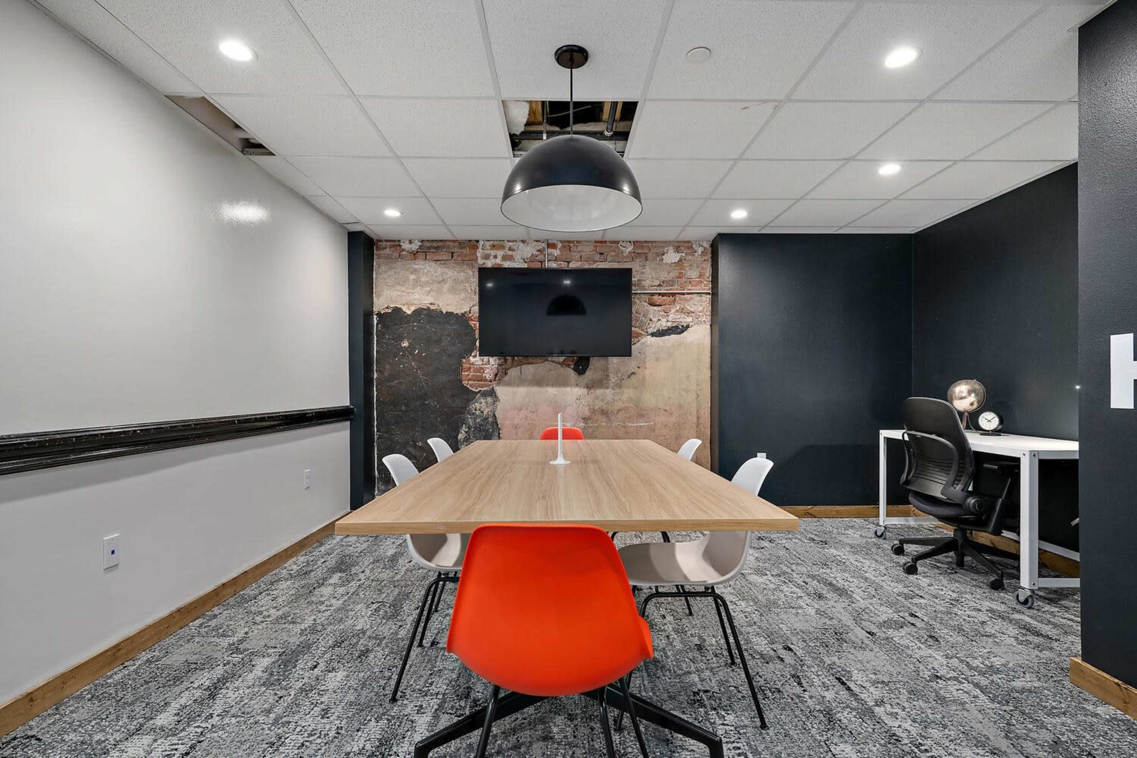 A modern meeting room features a large wooden table surrounded by white and orange chairs, with a black wall displaying a television and a section of exposed brick.