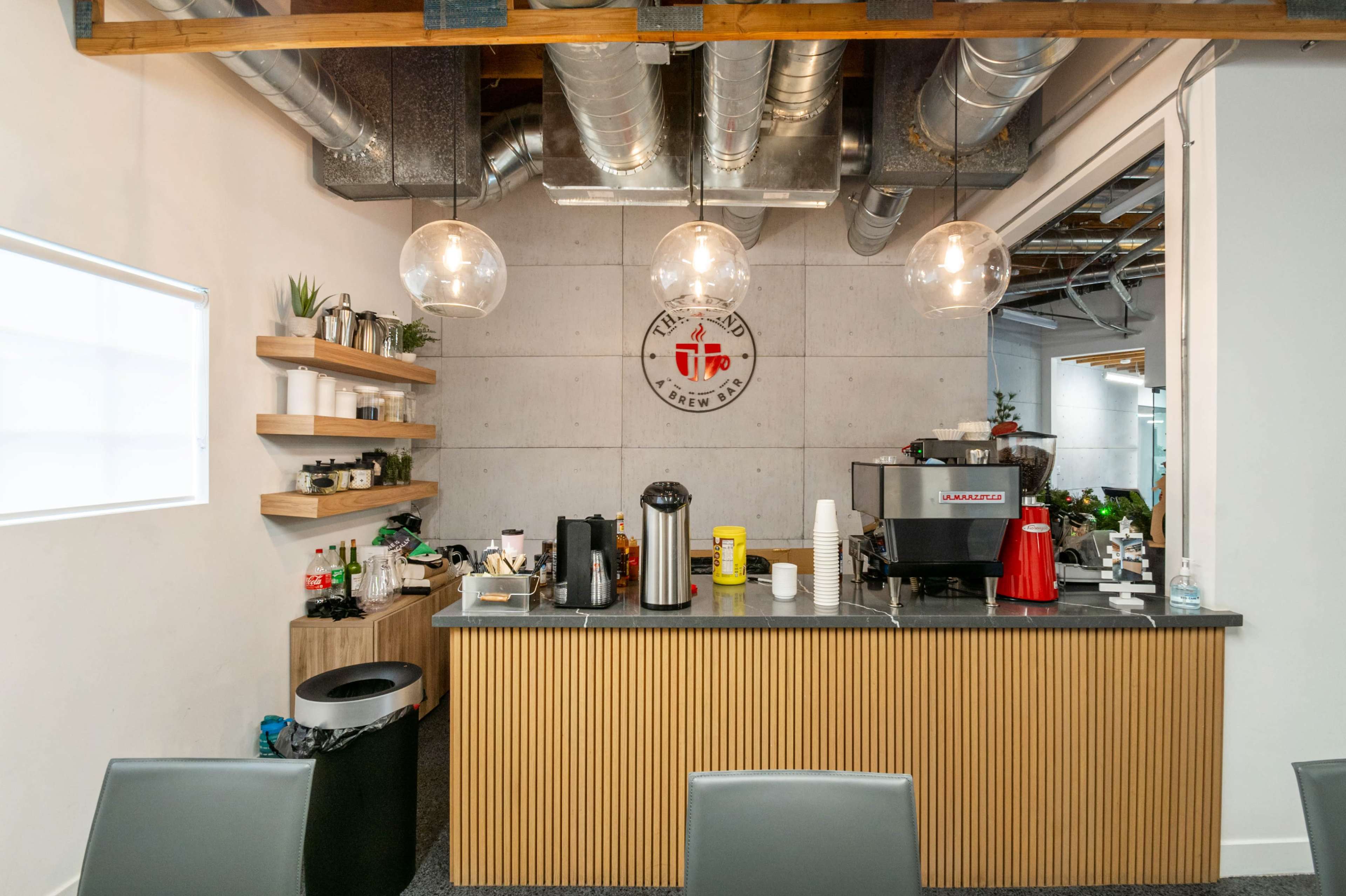 The image shows a modern coffee station with a wooden counter, shelves displaying plants and items, and pendant lighting overhead in a minimalist workspace.