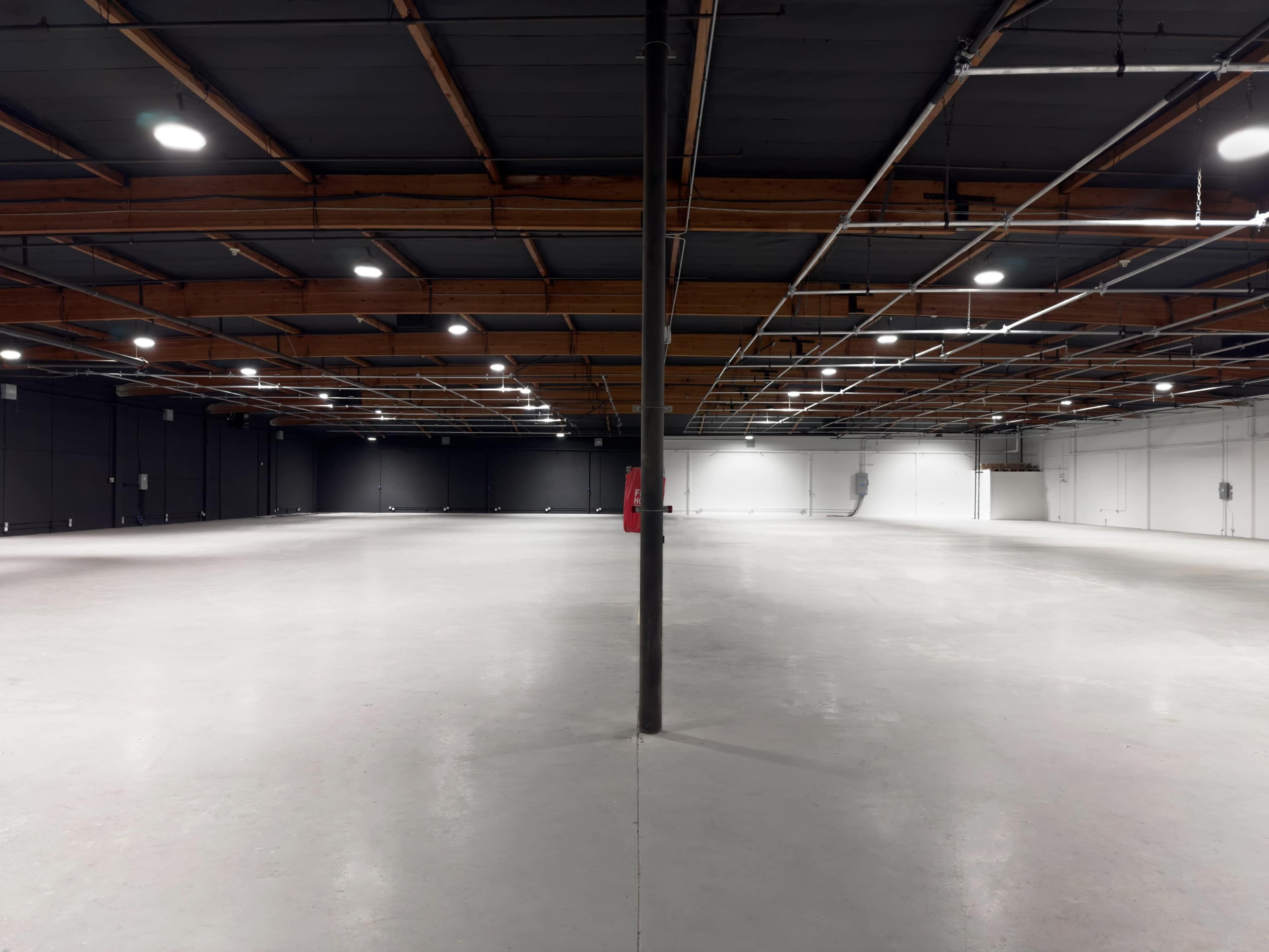 A large, empty warehouse space with concrete flooring, a visible central support pole, and evenly distributed overhead lighting.