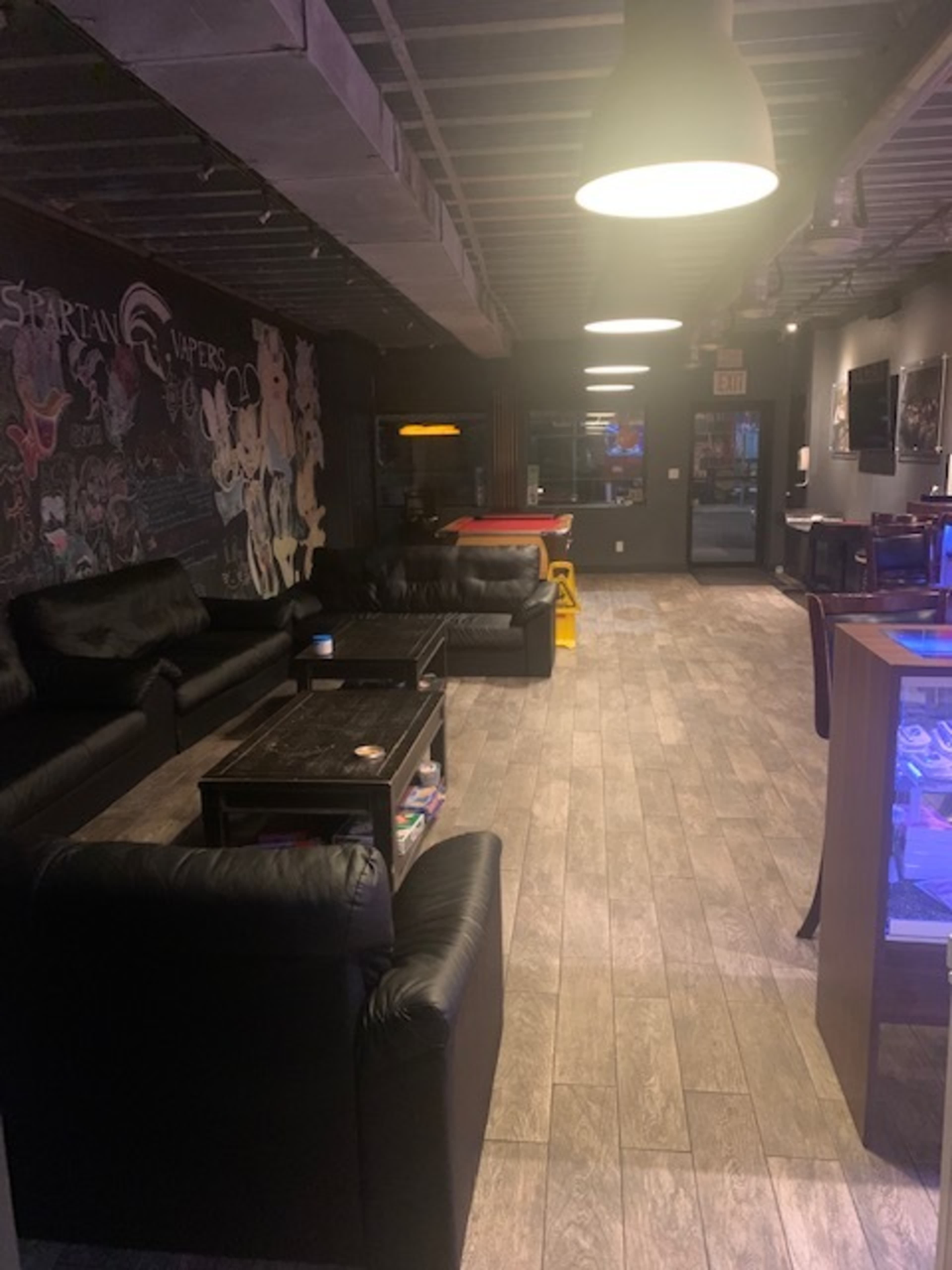 A lounge area with black leather sofas, wooden flooring, and a dimly lit environment featuring wall art and a gaming table.