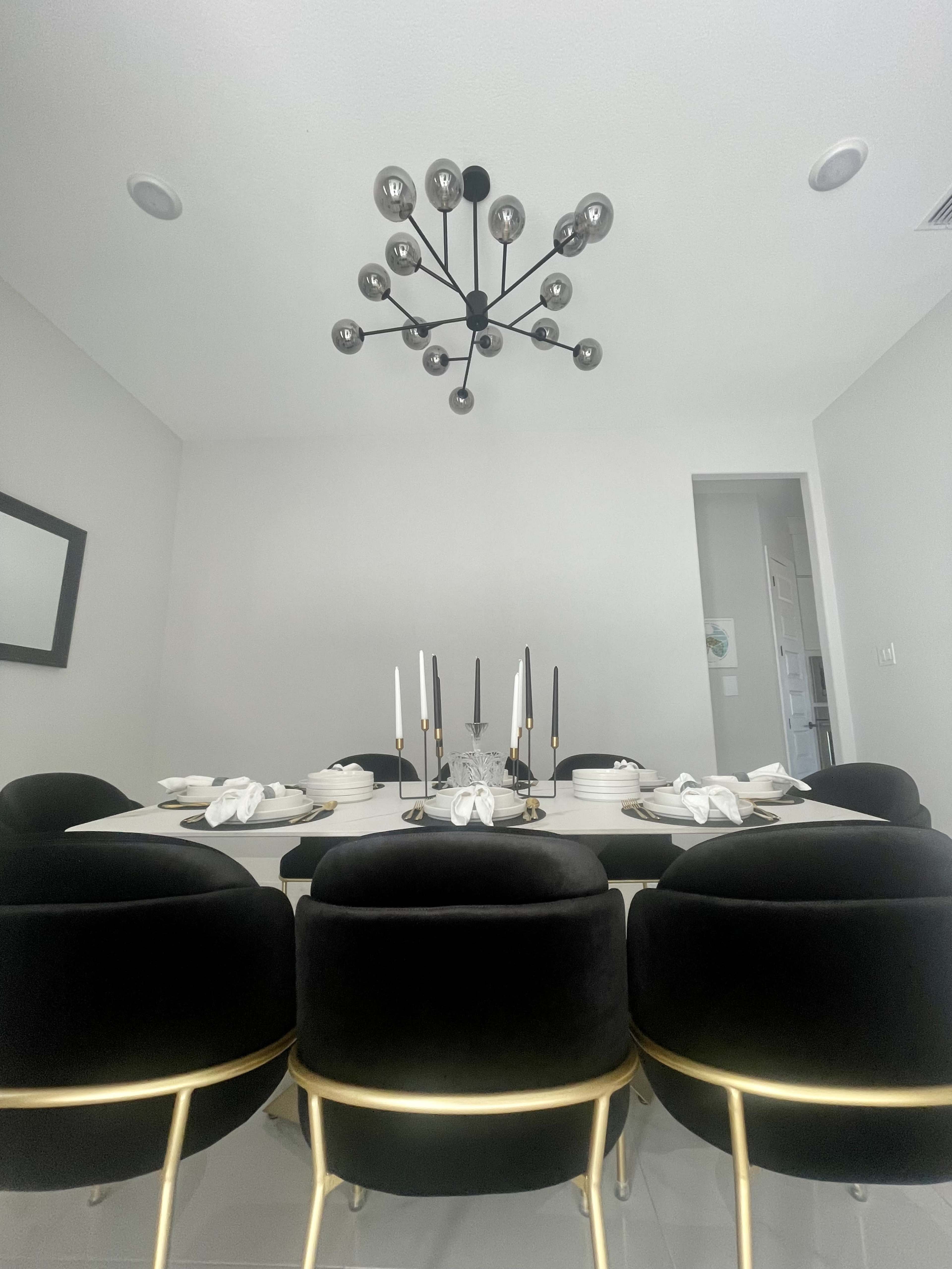 A modern dining room features a large table set with white plates and black and white candles, surrounded by black chairs with gold accents and a decorative light fixture overhead.