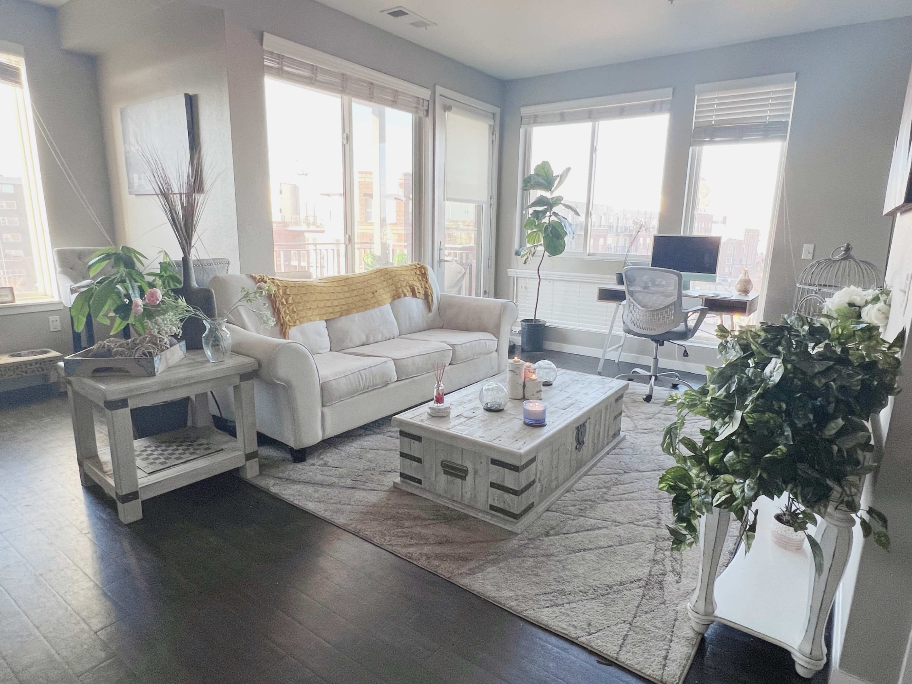 A bright living room with a white couch, a wooden coffee table, potted plants, and a desk with a chair near large windows.