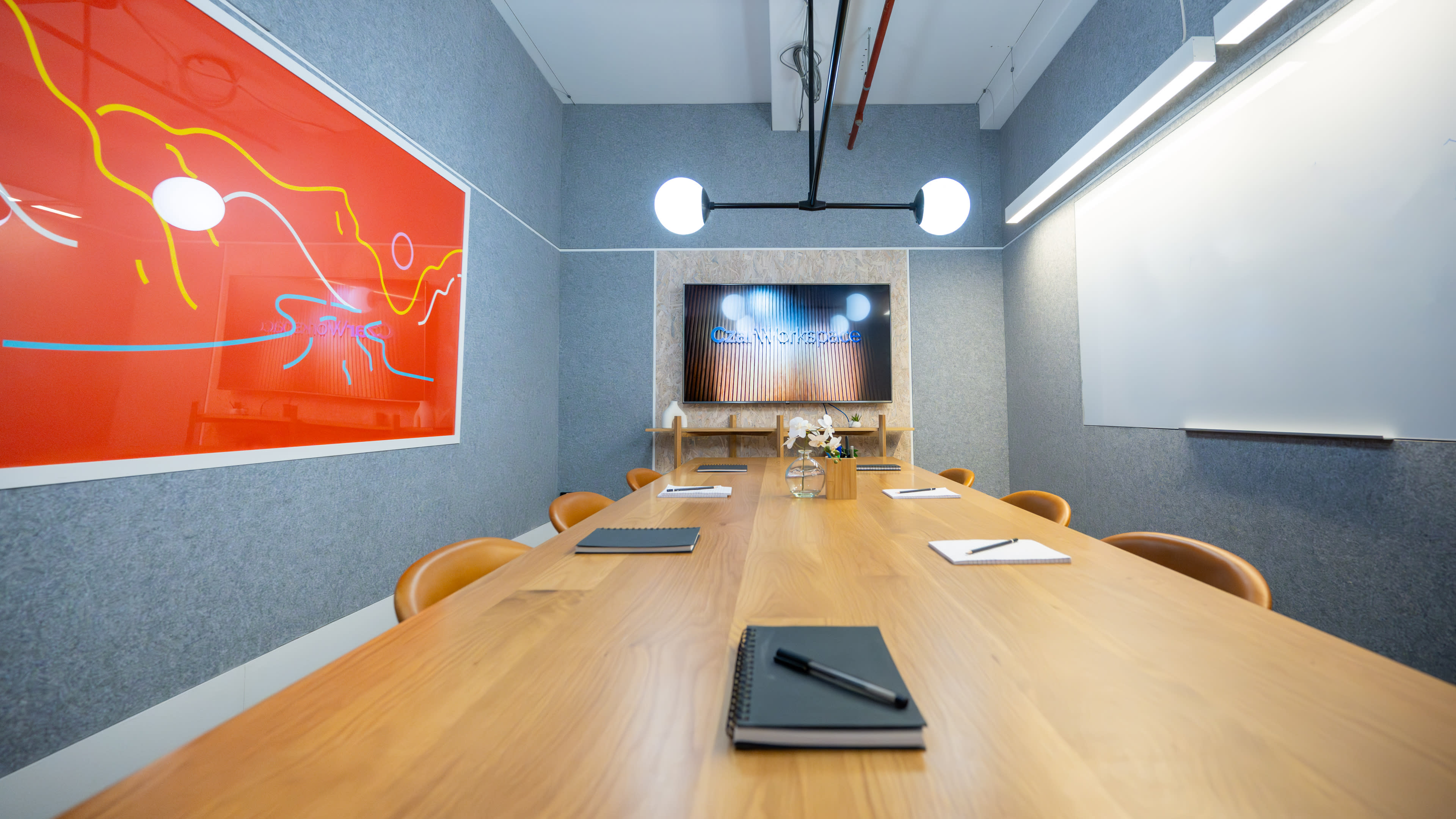 A modern conference room features a long wooden table with notepads and pens, a large screen on the wall, and colorful wall art.