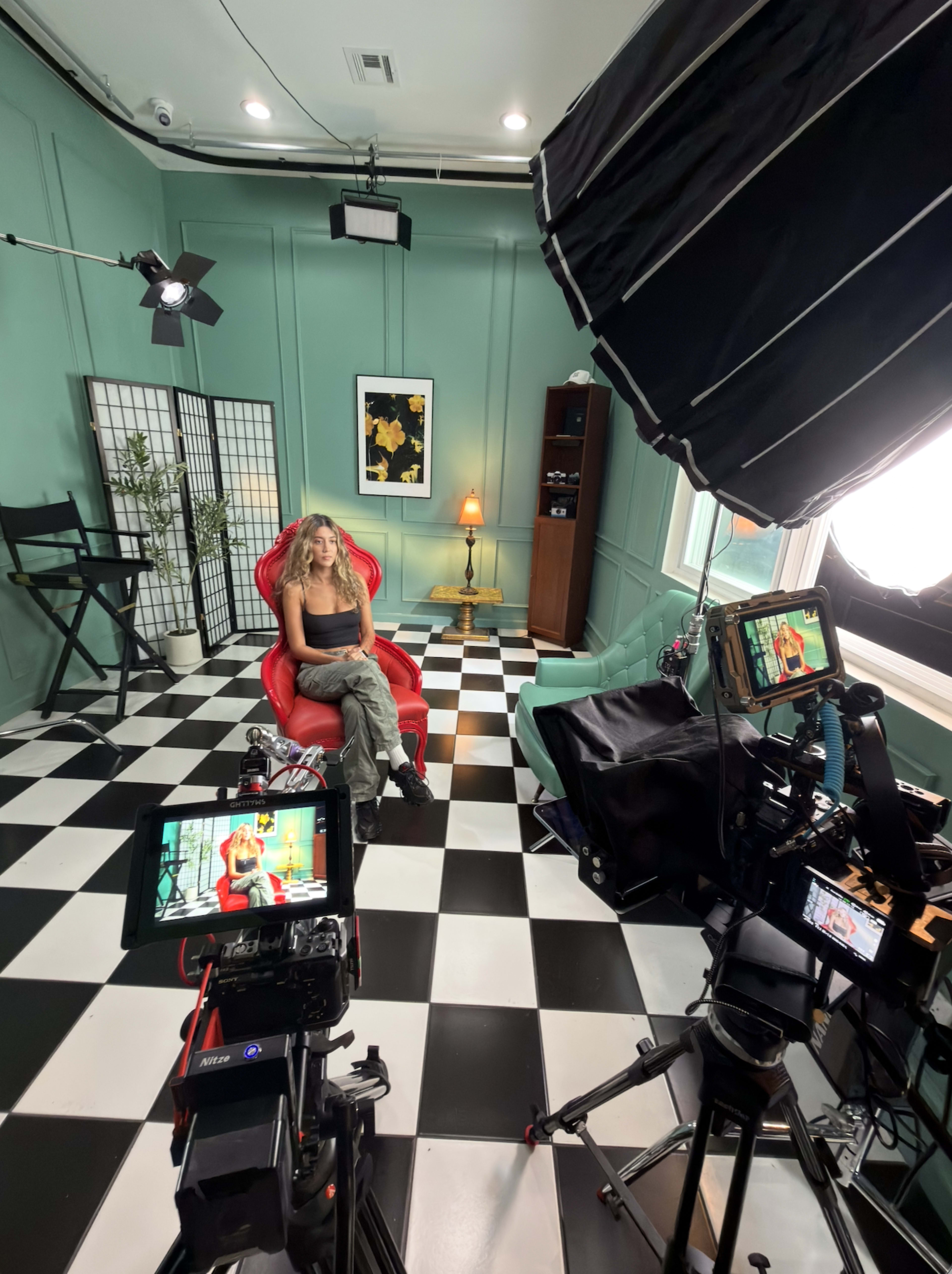 A woman sits in a red bean bag chair in a room with green walls and a checkered floor, surrounded by filming equipment and lights.