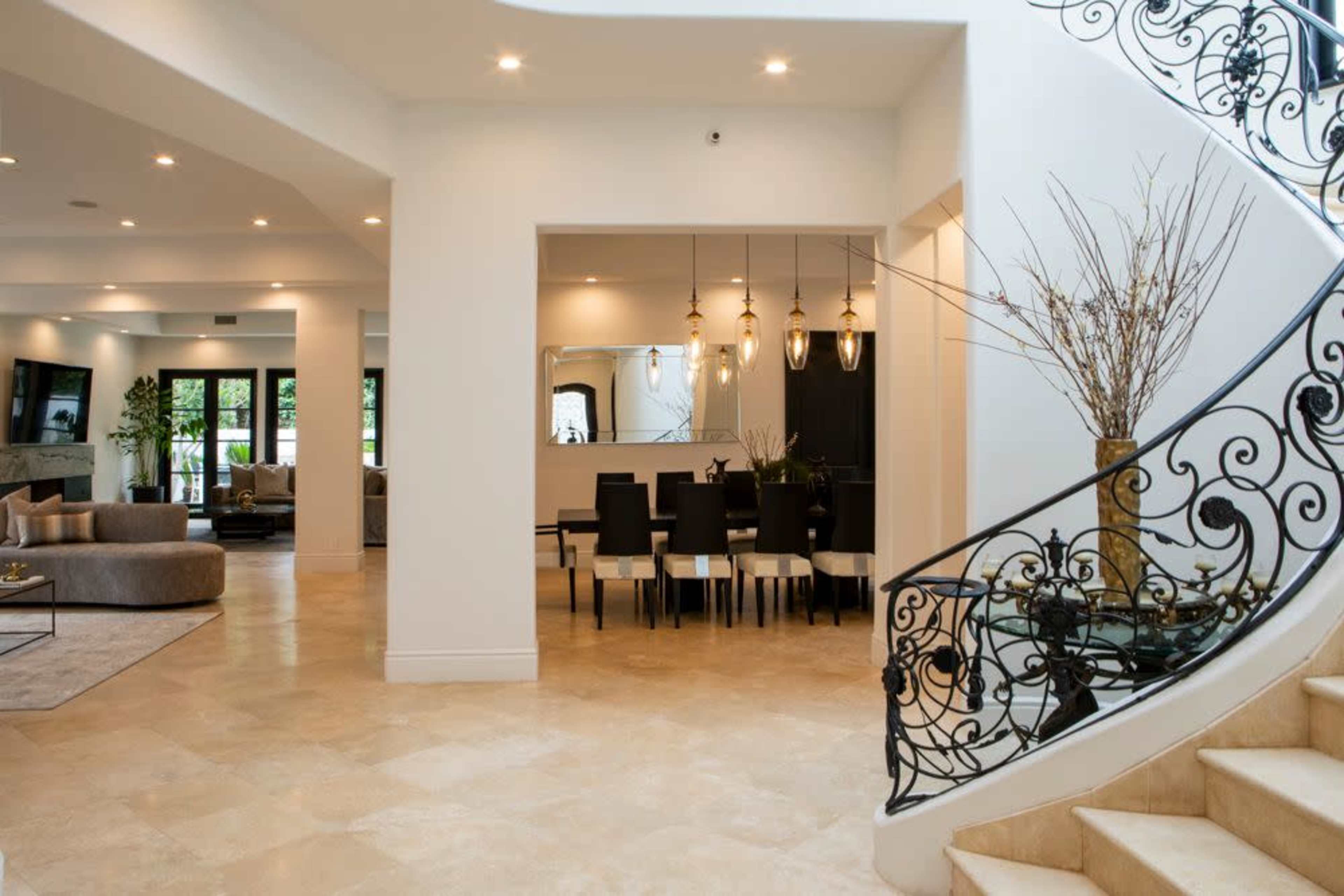 A spacious interior featuring a staircase with ornate railings, leading to a dining area with a table and chairs, illuminated by pendant lights.