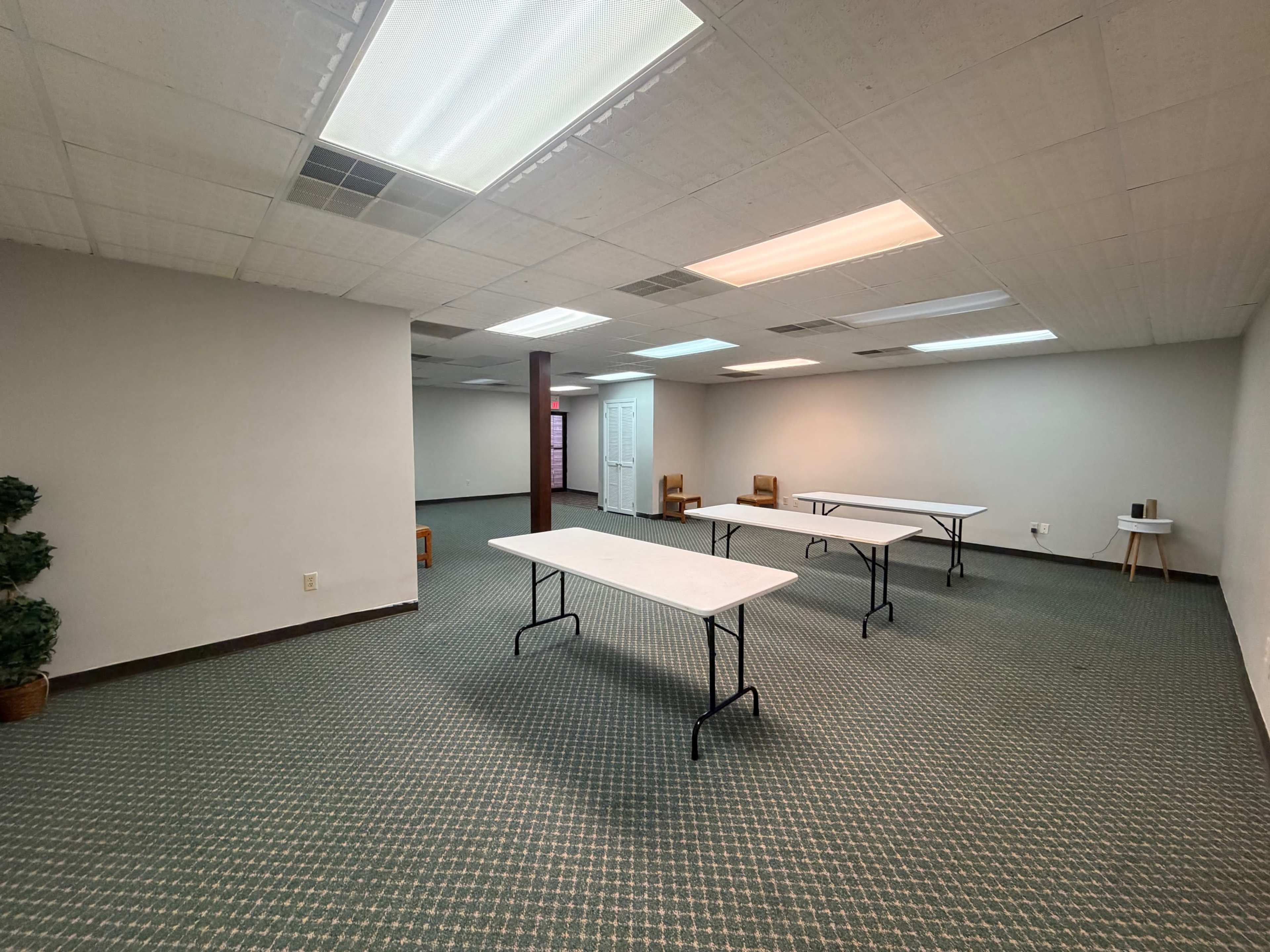 The image shows a spacious, empty room furnished with several folding tables and a few chairs, all set against a carpeted floor and illuminated by ceiling lights.