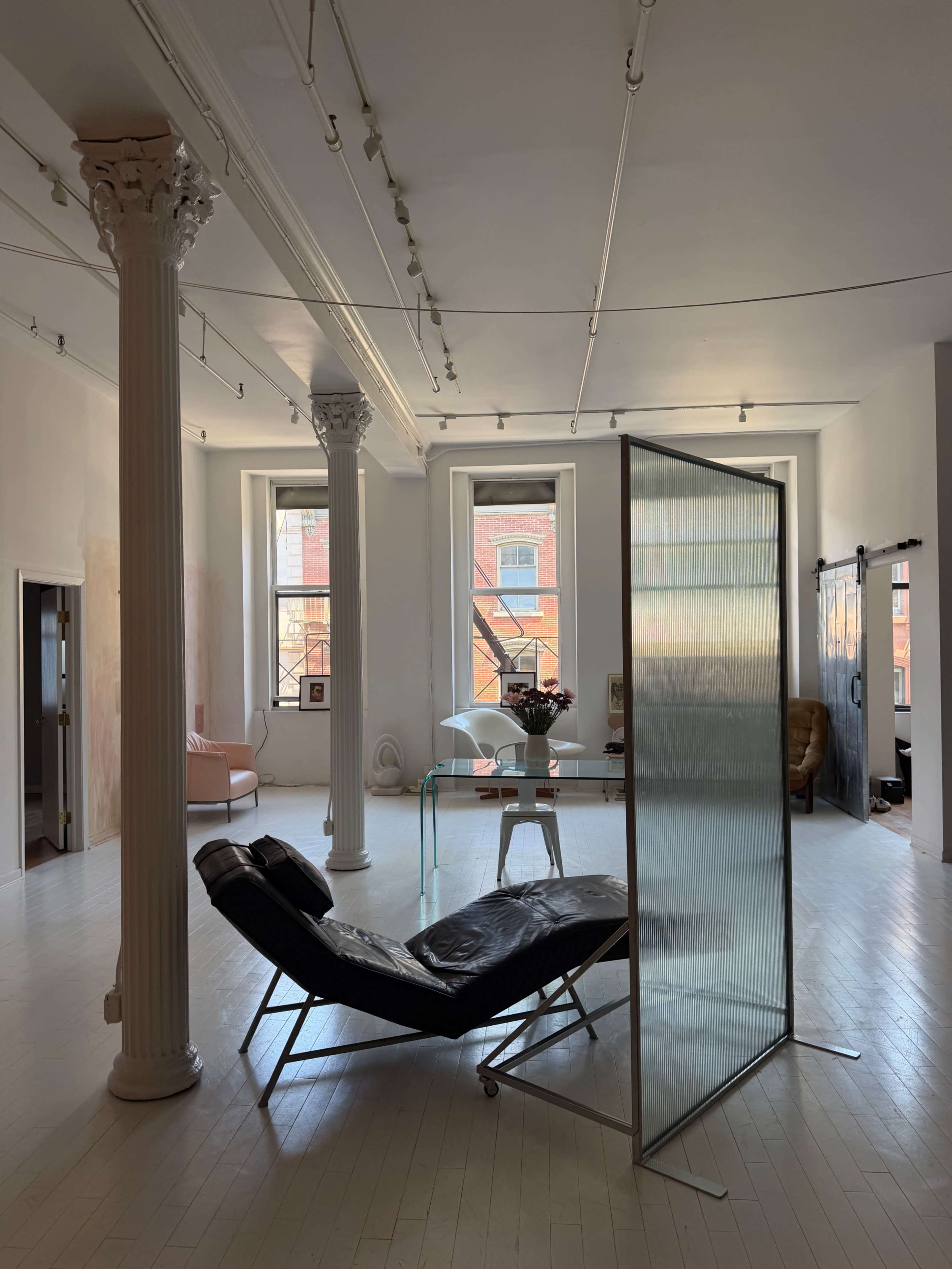 The image shows a spacious, minimalist room with large windows, featuring a black lounge chair, a transparent divider, and decorative elements including a glass table and potted plants.