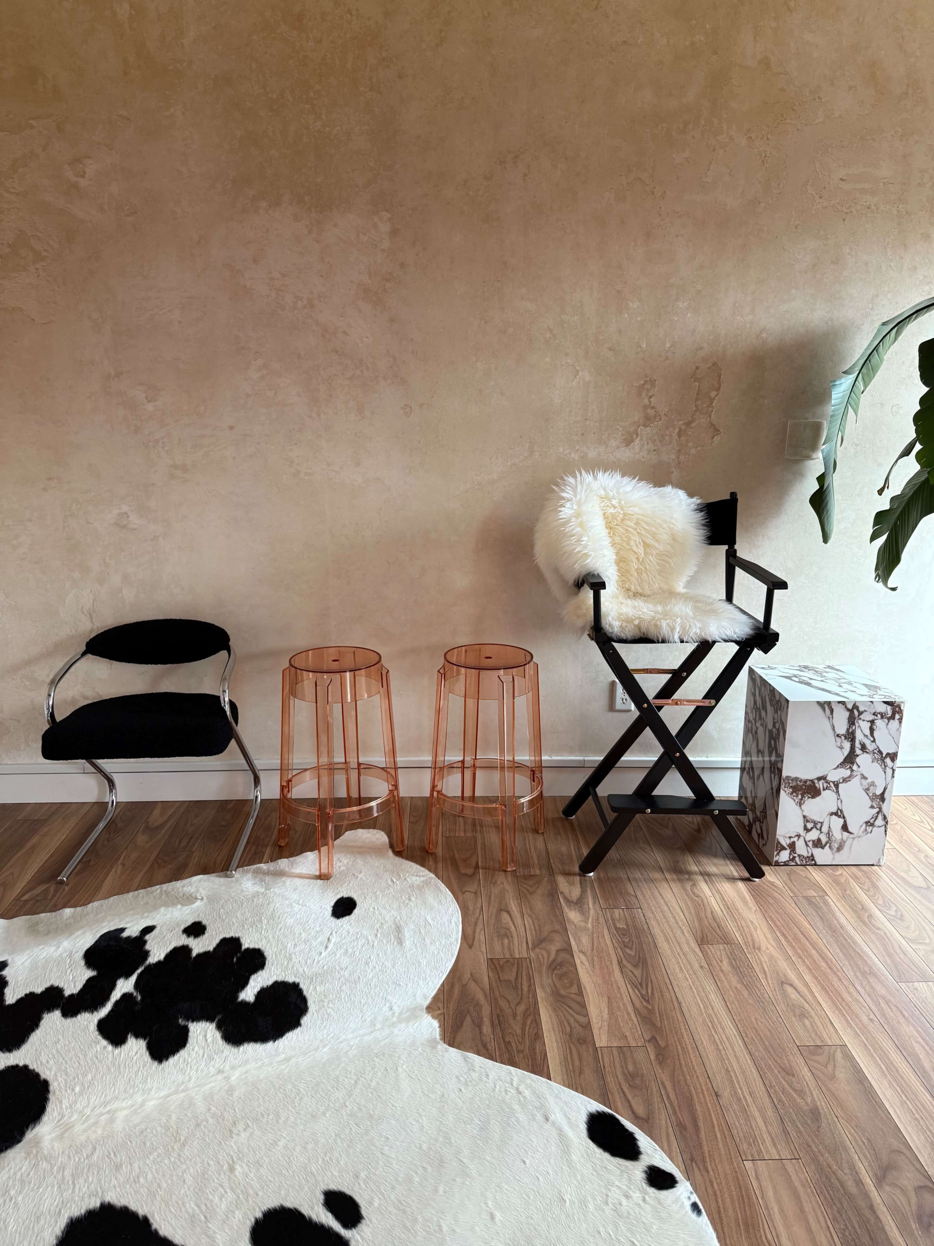 The image shows two transparent orange stools, a black director's chair with a white fur throw, a black chair, and a marble-patterned side table against a textured beige wall.