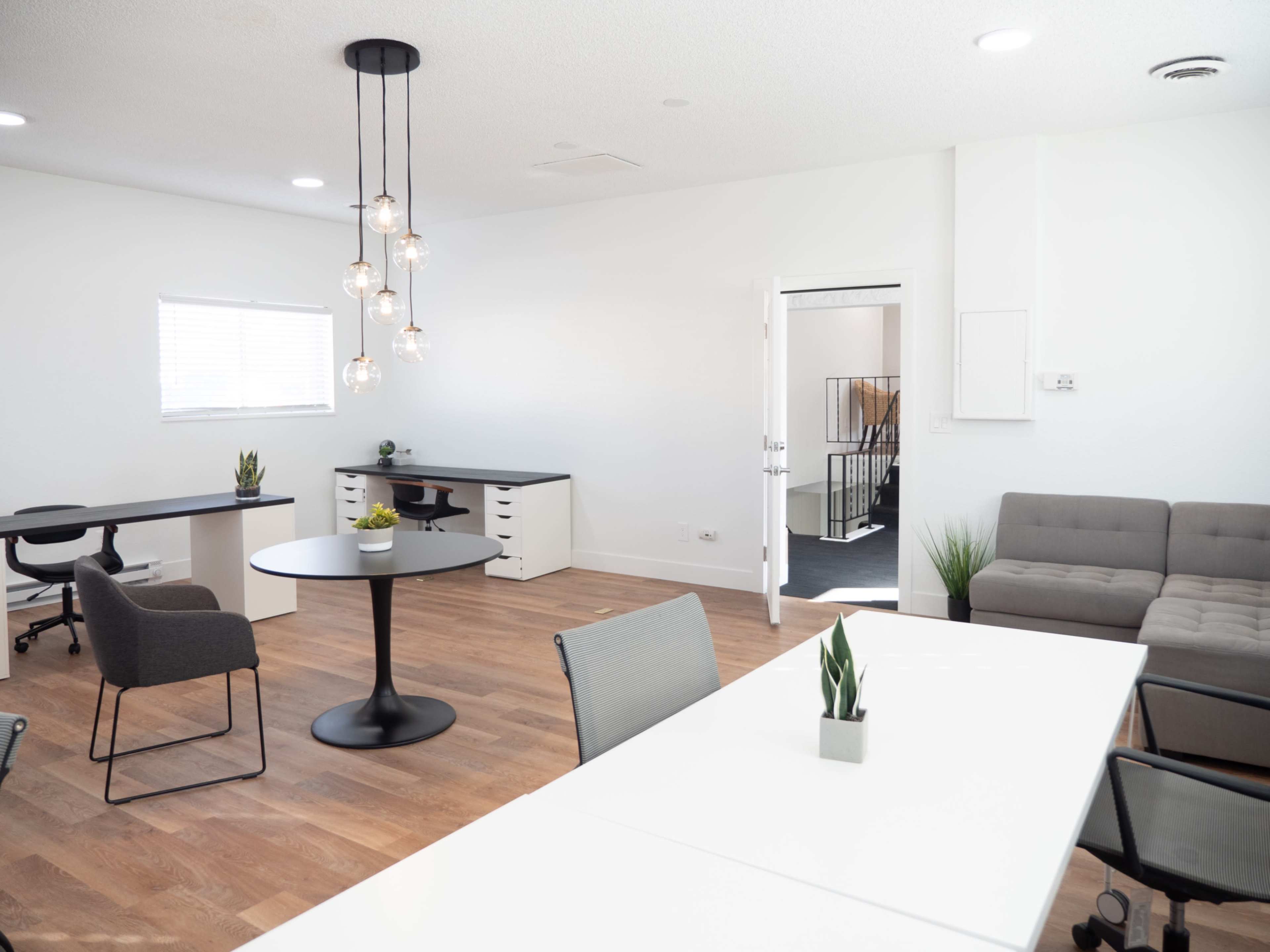 The image shows a modern office space with two desks, a round table with chairs, and a seating area, all featuring minimalist decor and wooden flooring.