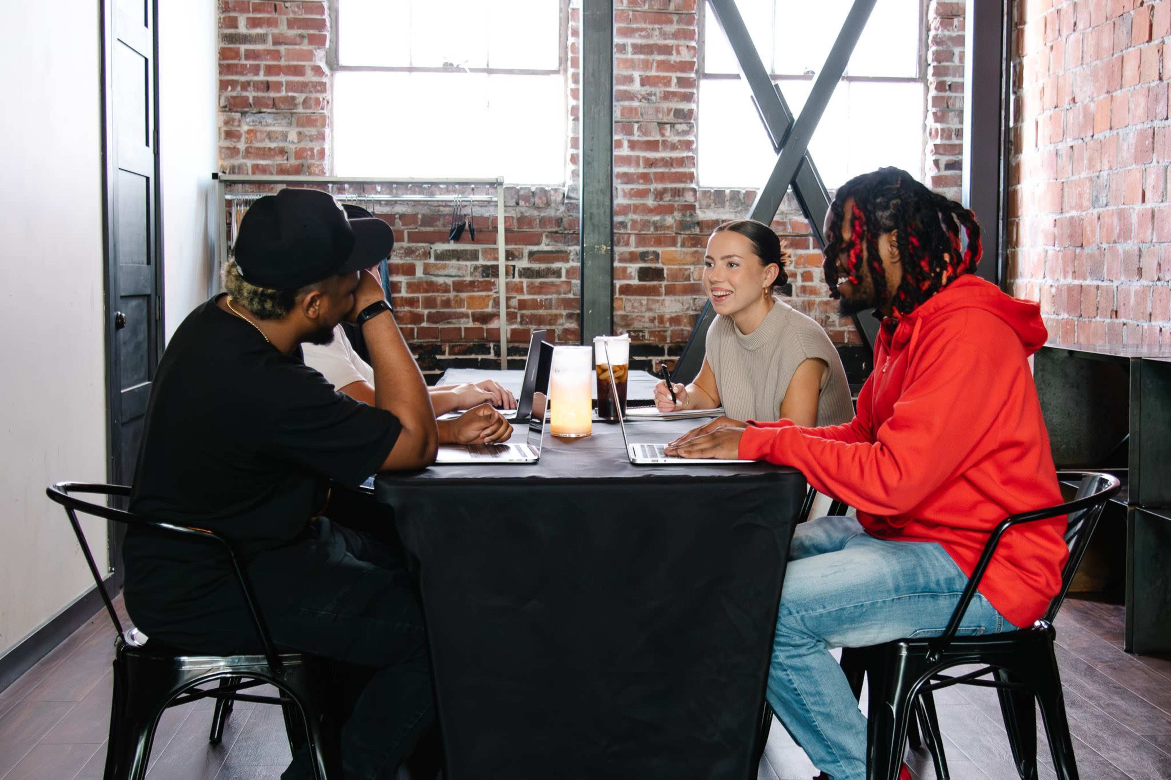 A group of four people sits around a table in a brick-walled room, engaged in a discussion while working on laptops.