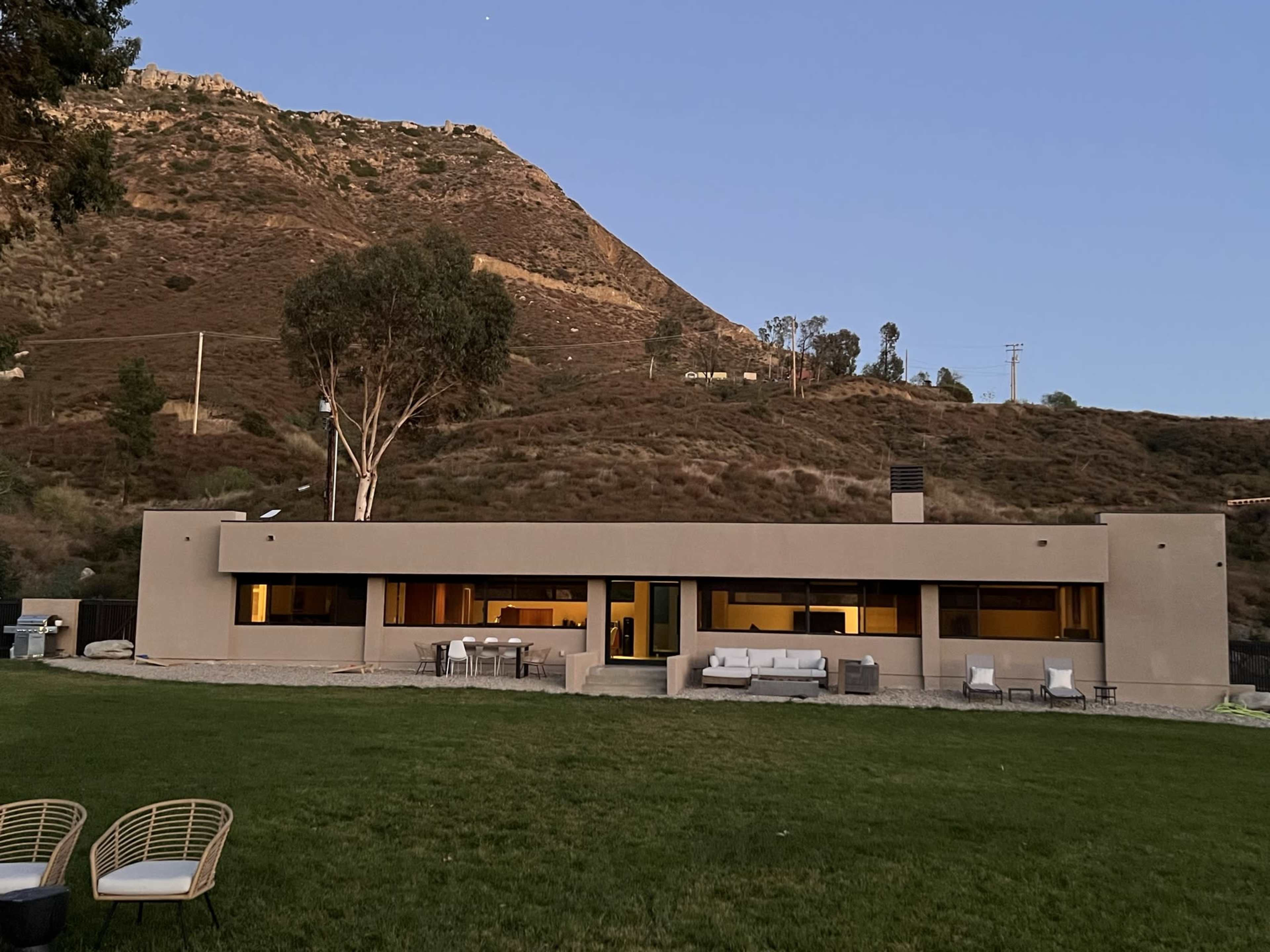 A modern single-story house situated on a grassy area with a hillside in the background at dusk.