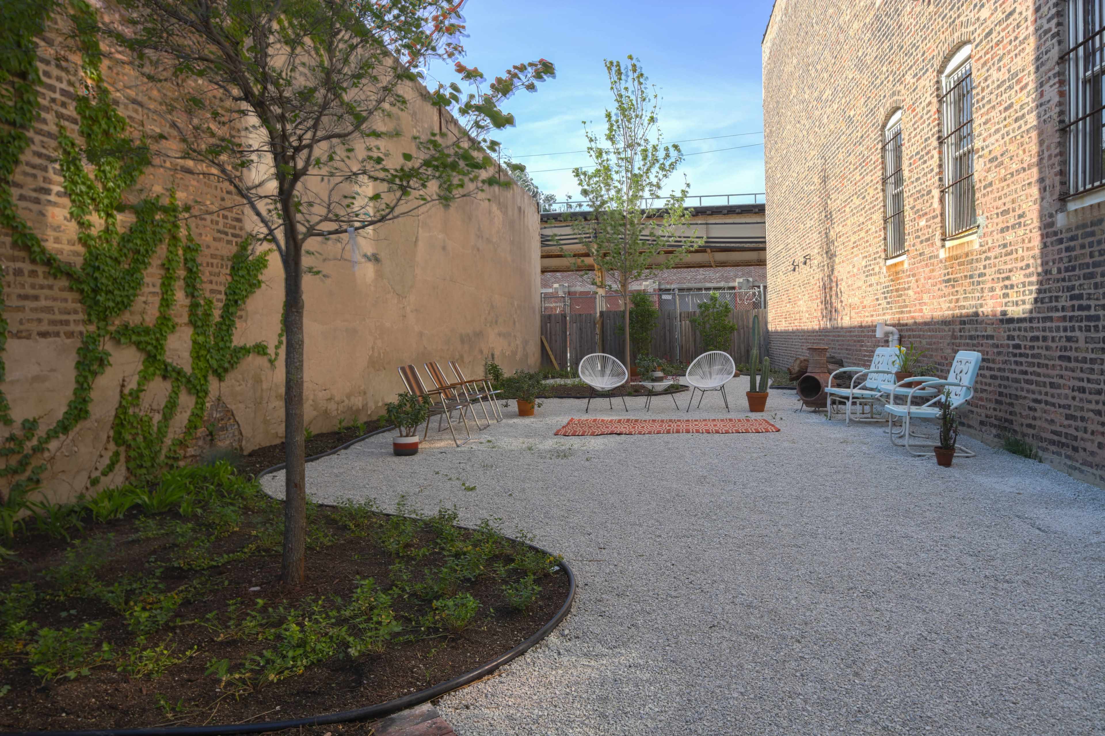 A narrow outdoor space with gravel flooring, featuring seating arrangements, potted plants, and a small area rug surrounded by brick walls.
