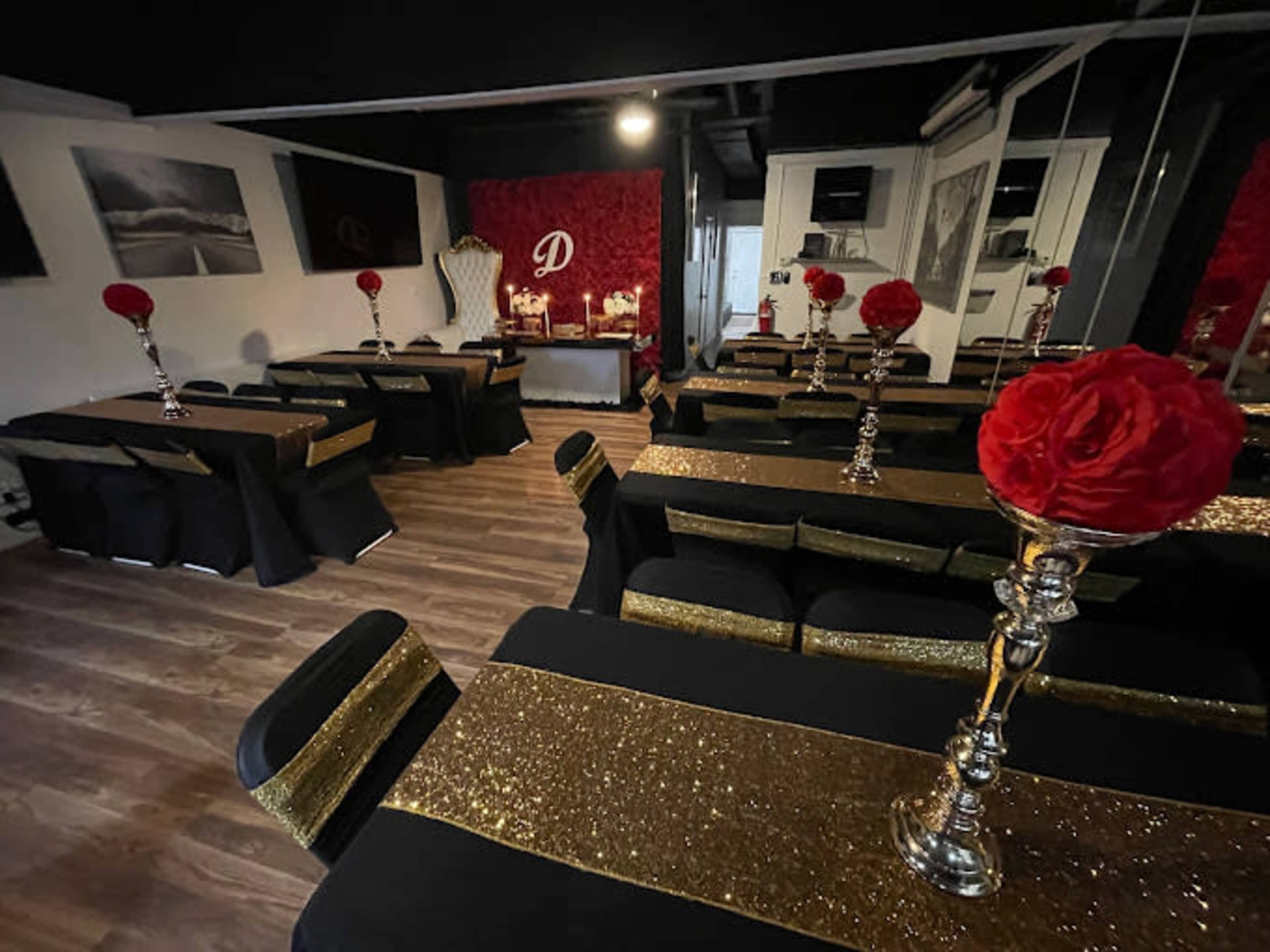 The image shows a decorated event space with multiple tables covered in black and gold tablecloths, each adorned with candlestick centerpieces and red floral arrangements.