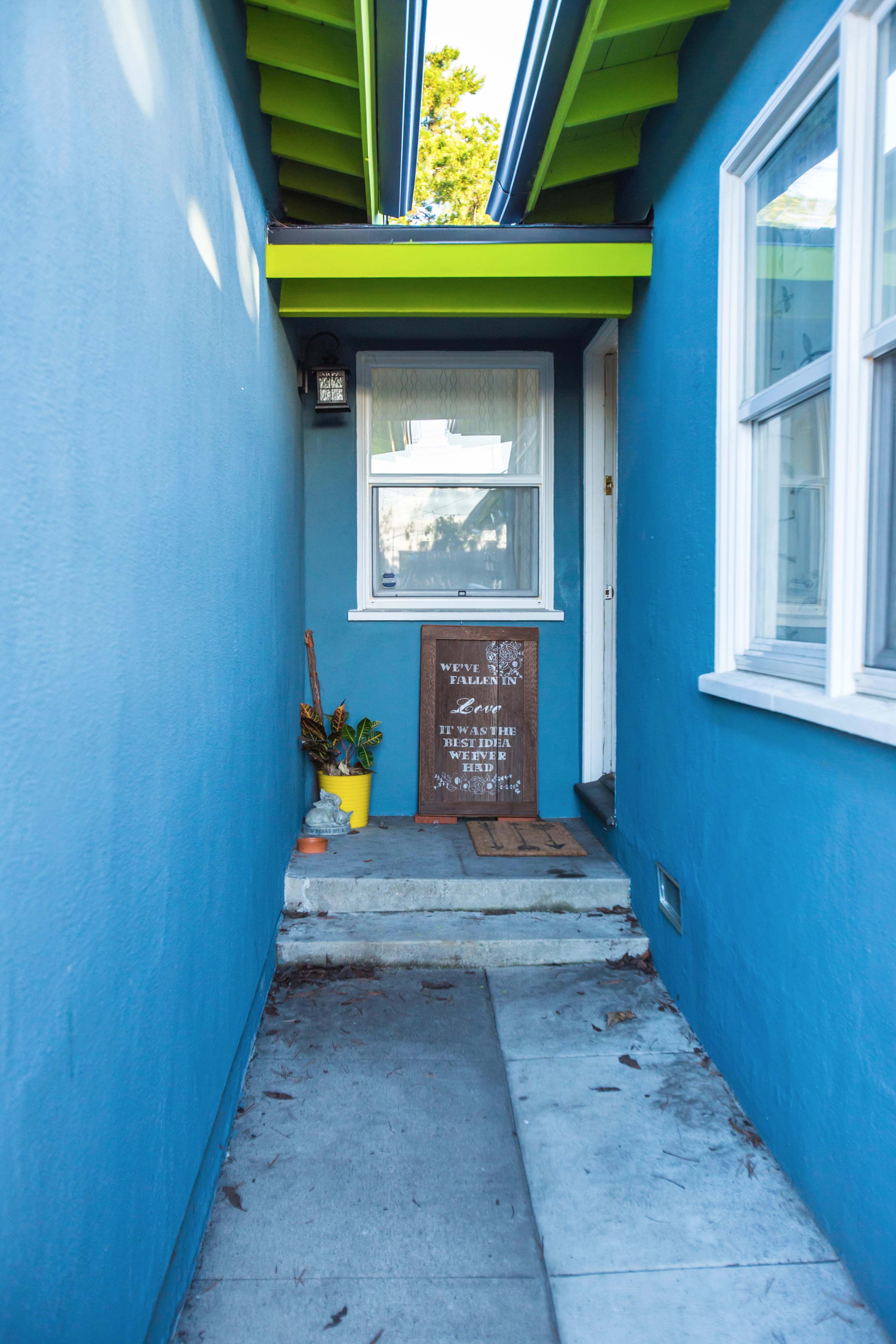 A narrow walkway leads to a blue house entrance, featuring a decorative sign and a potted plant beside the door.