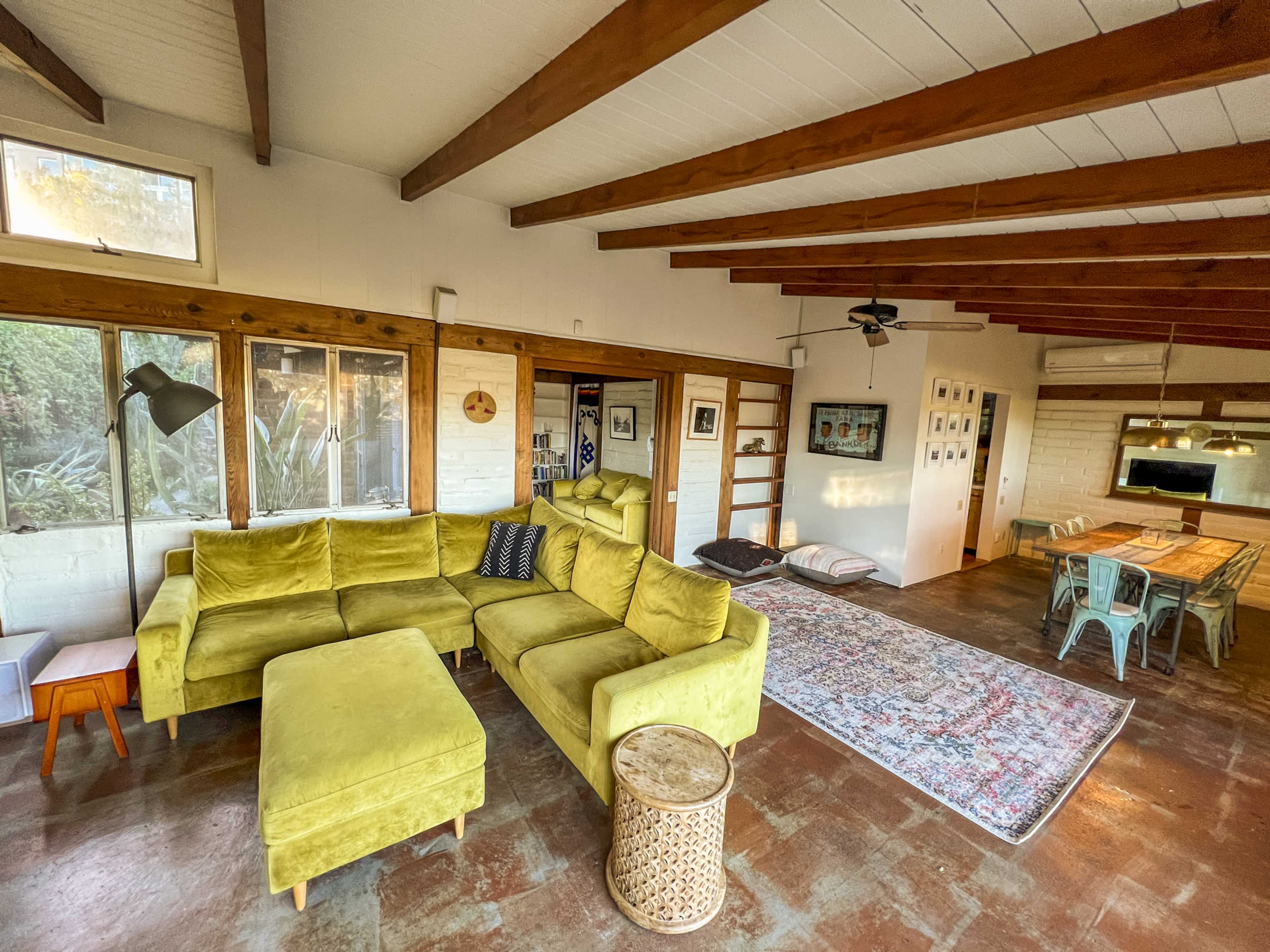 A spacious living area features a yellow sectional sofa, a wooden dining table, and exposed wooden beams on the ceiling.