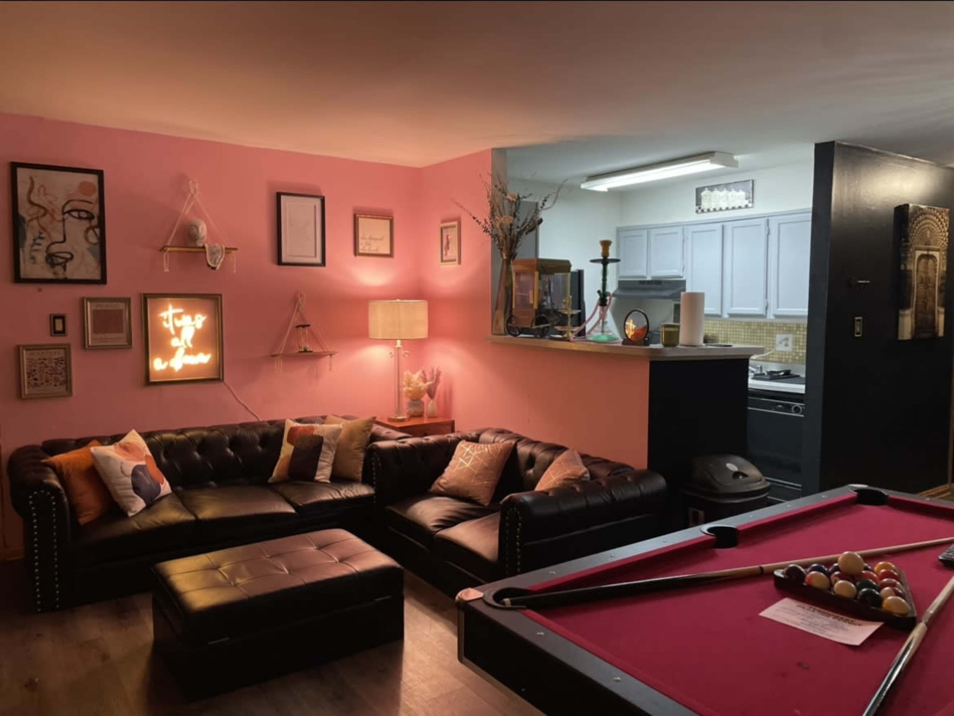 A cozy room featuring a black leather couch with colorful pillows, a pink wall, and a pool table in the foreground, while a kitchen area is visible in the background.