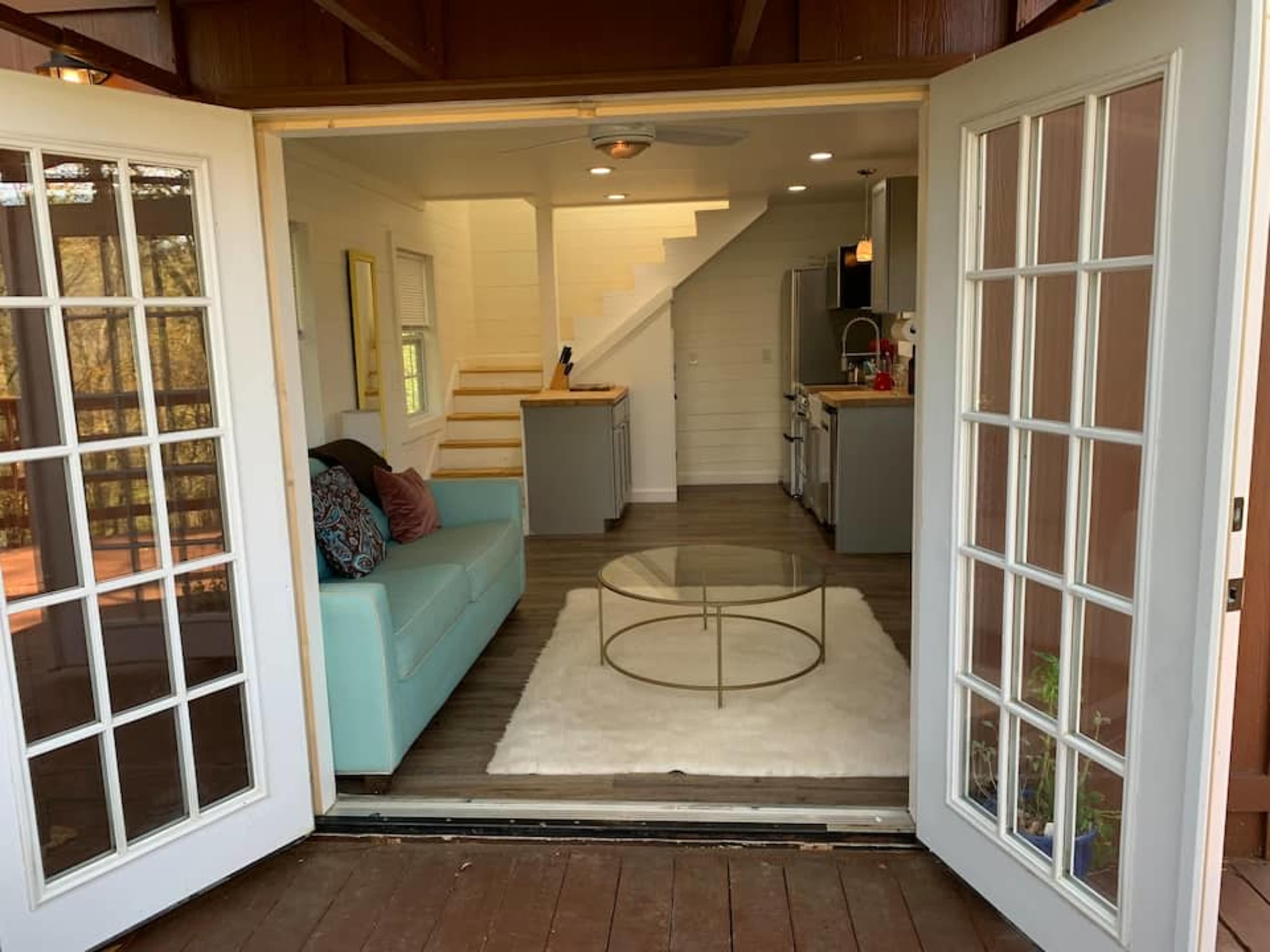 A set of French doors opens to a living area featuring a light blue couch, a round glass coffee table, and a staircase leading to an upper level.