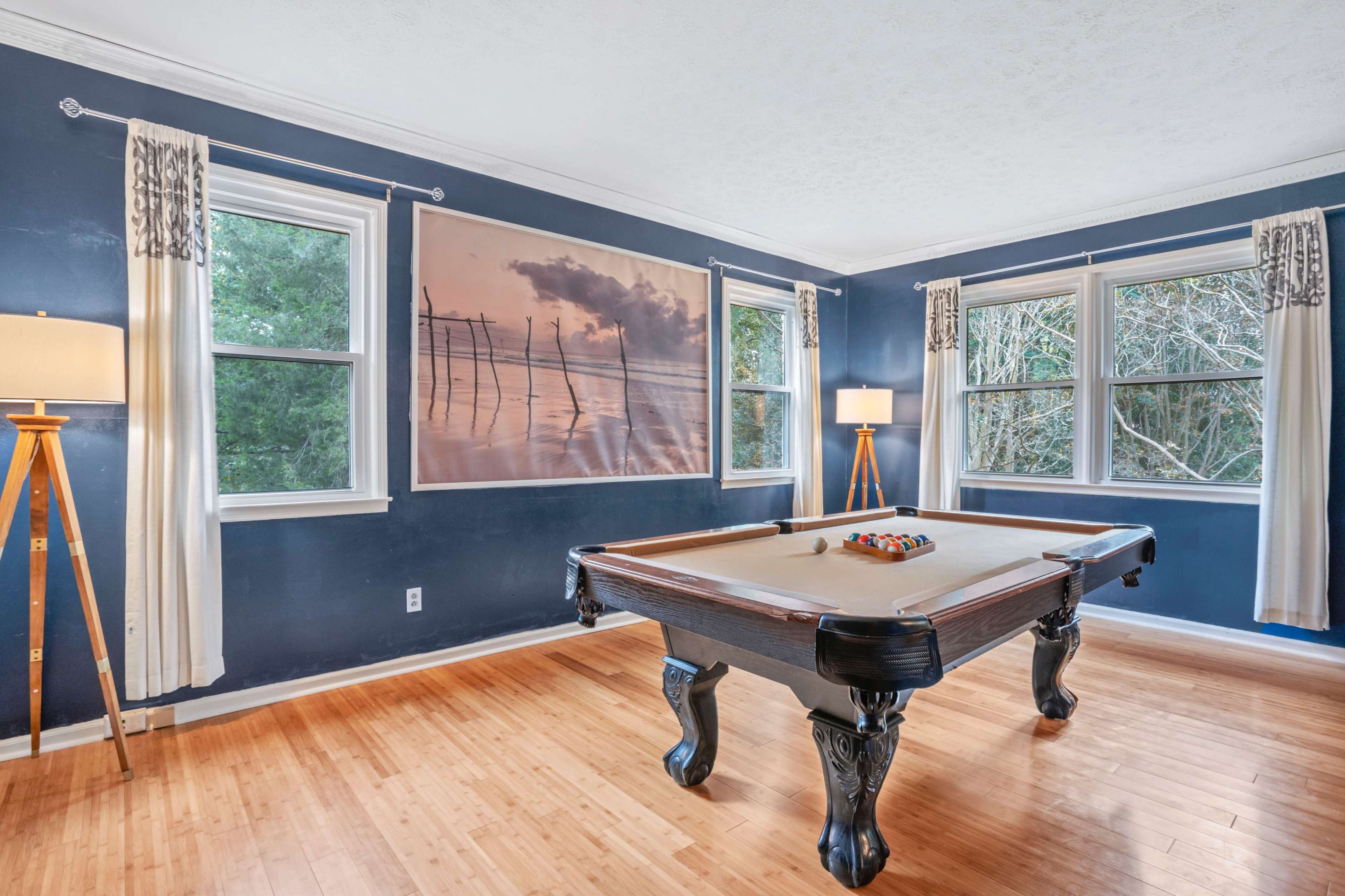The room features a pool table centered beneath large windows with a scenic photograph on the wall and two lamps on either side.