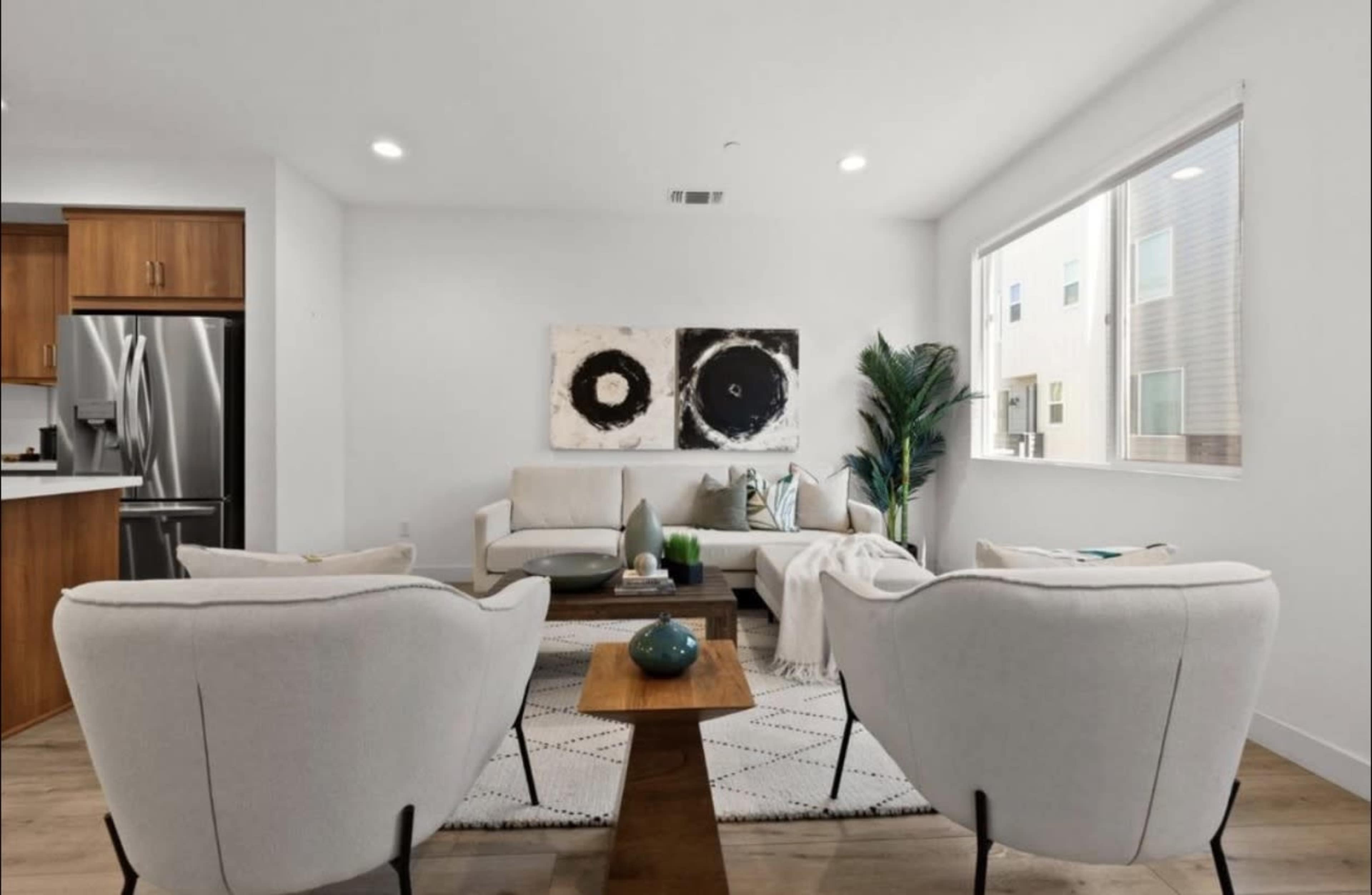 The image shows a modern living room with a neutral color palette, featuring two armchairs and a sofa arranged around a wooden coffee table, along with art on the wall and a plant in the corner.