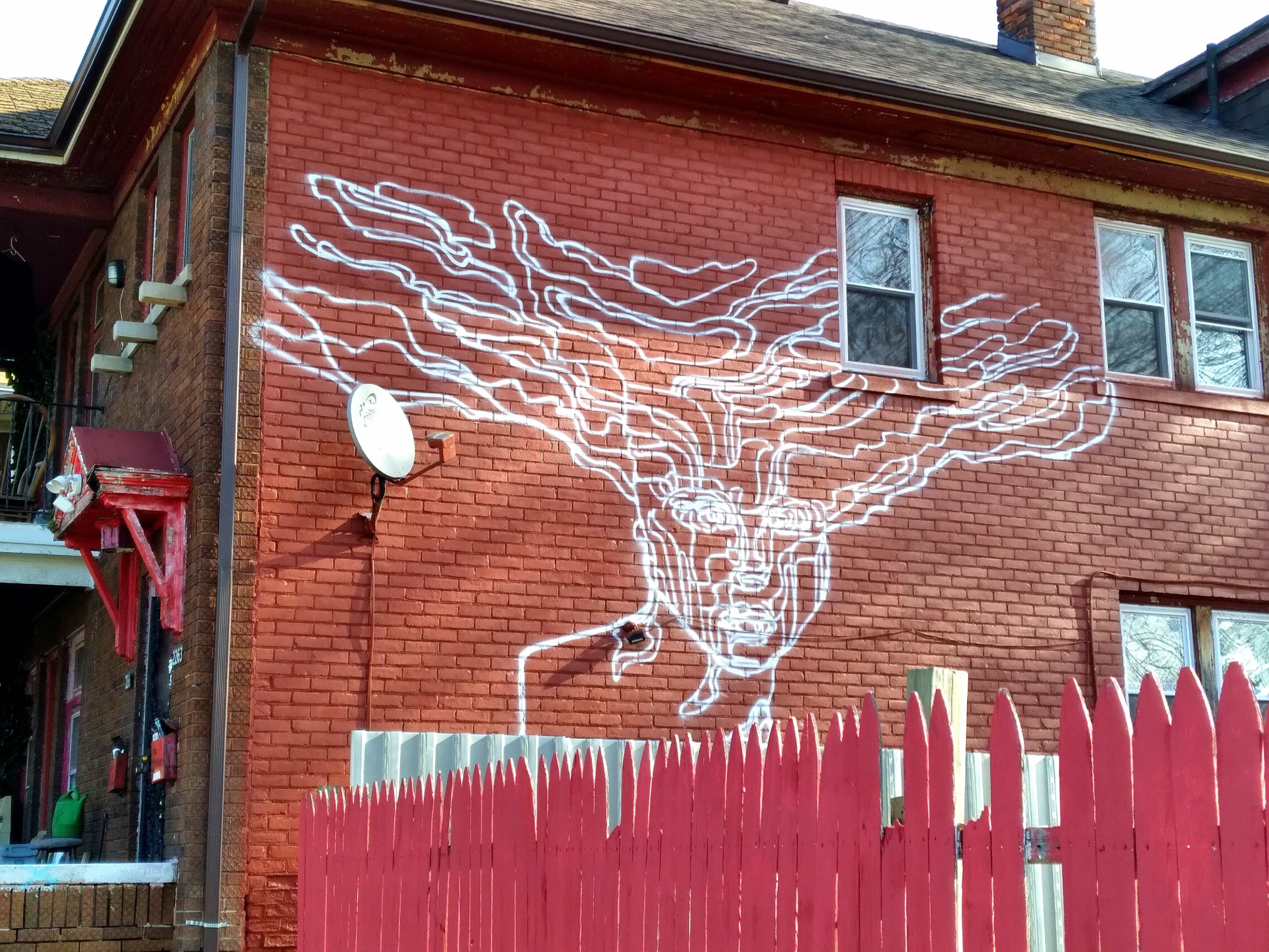A large white mural depicting a face with flowing hair is painted on a red-brick building alongside a red picket fence.
