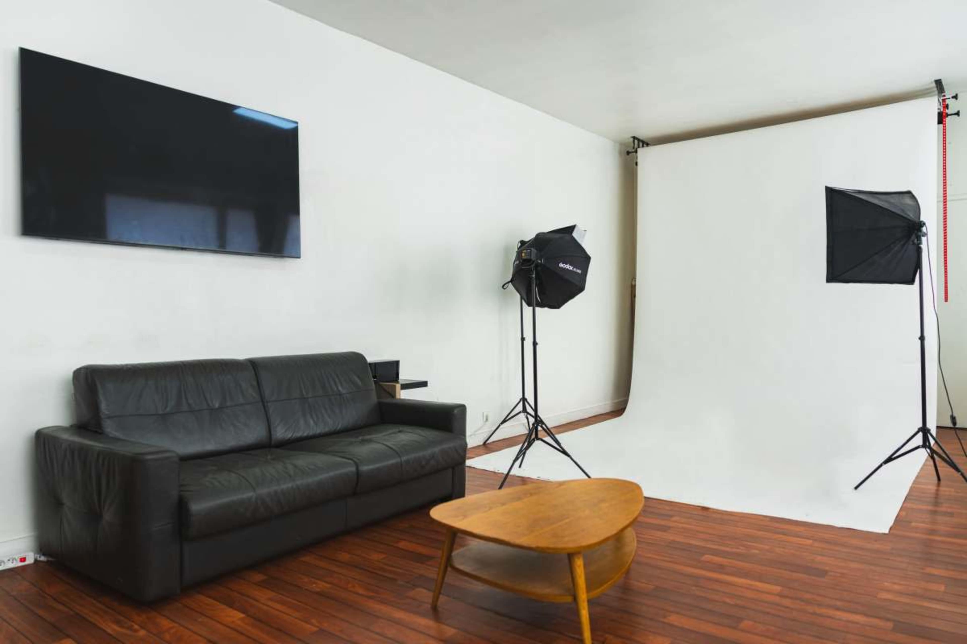 The image shows a photography studio with a black leather sofa, a wooden coffee table, and two light stands positioned near a white backdrop.