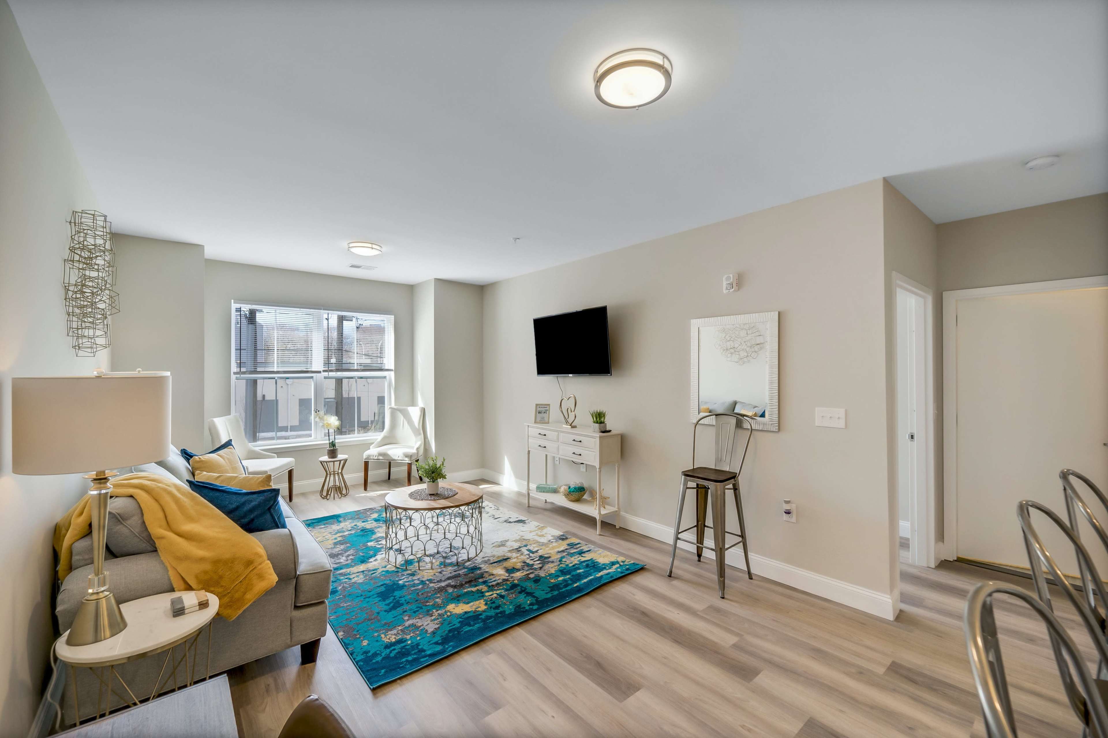 A modern living room with a light-colored sofa, a coffee table on a patterned rug, and a wall-mounted TV, along with a small dining area visible in the background.