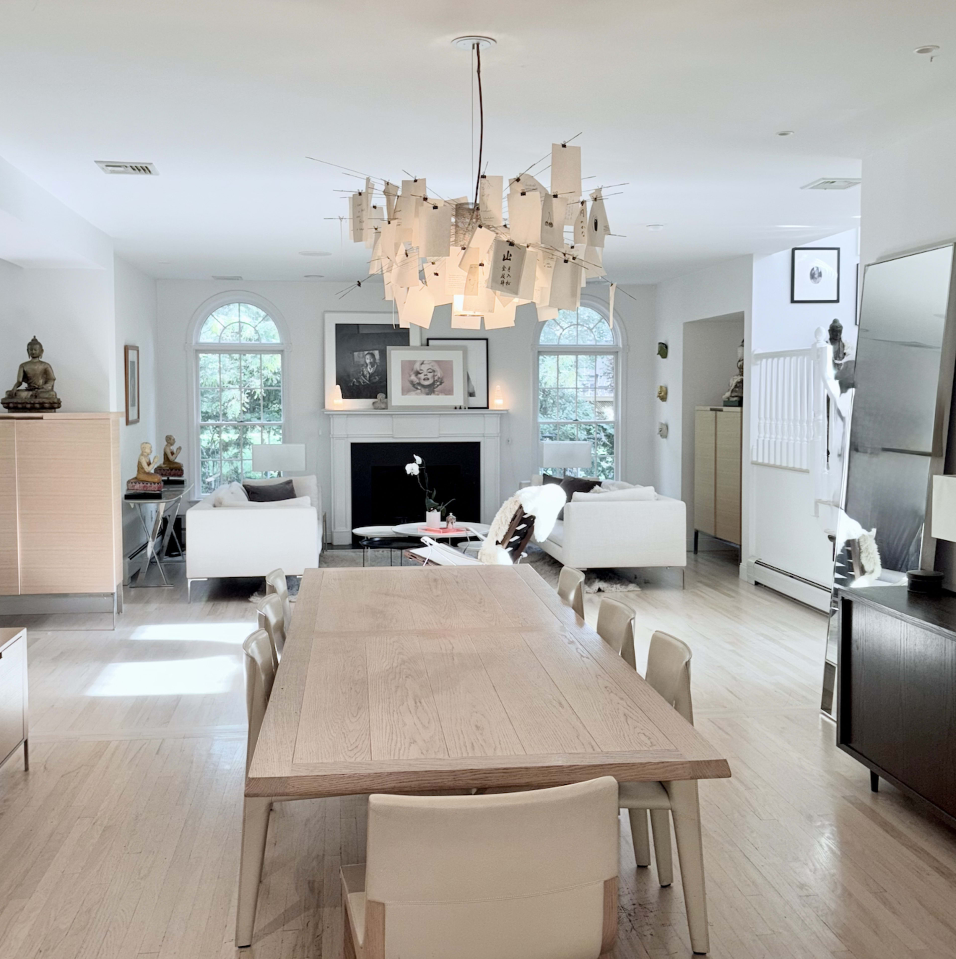 The image shows a spacious dining area with a large wooden table, surrounded by minimalist chairs, and a unique chandelier suspended above, while the room features two white sofas and a decorative fireplace.