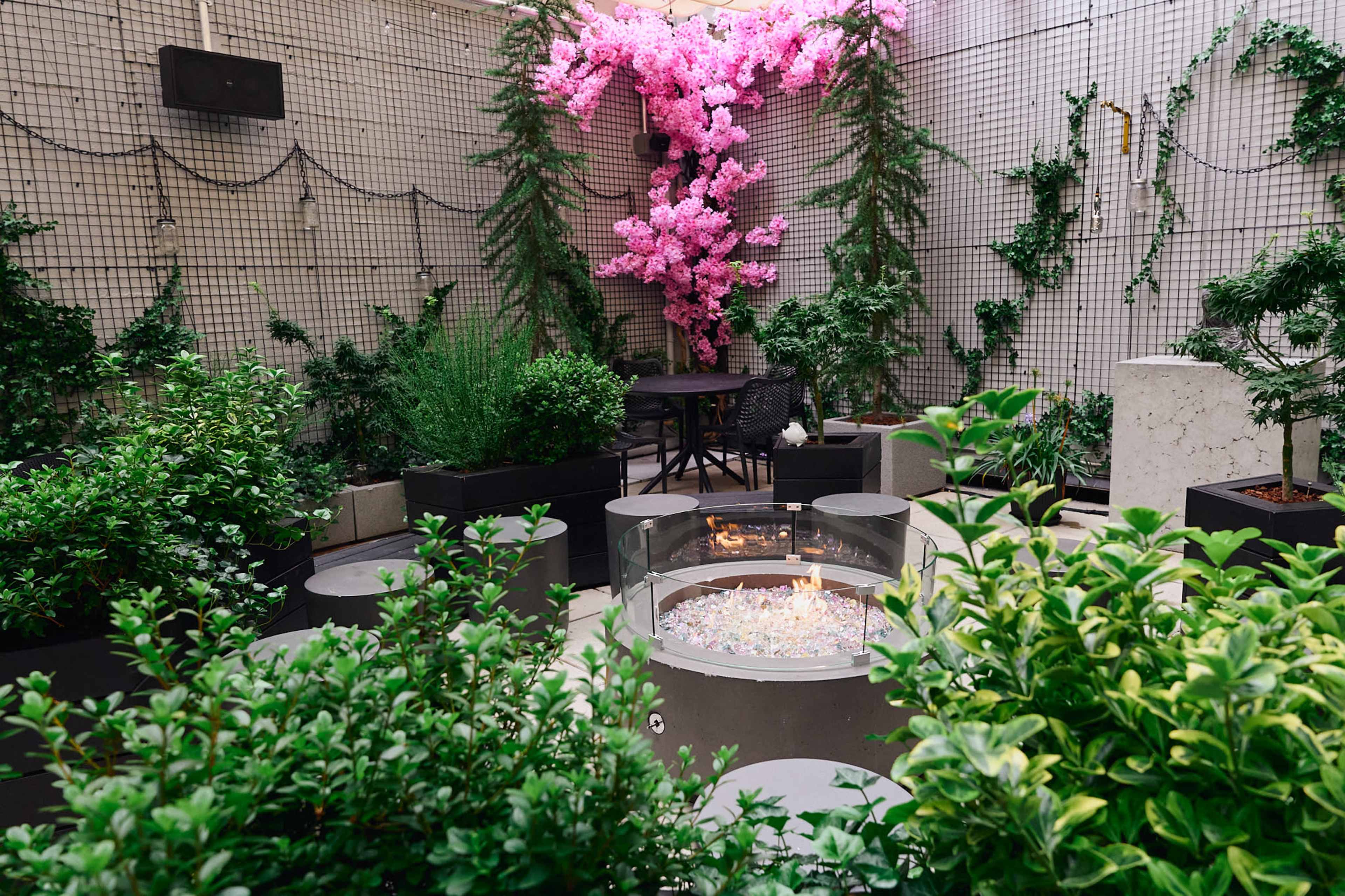 The image shows a serene courtyard with a central fire pit surrounded by lush greenery and a large pink flowering tree.