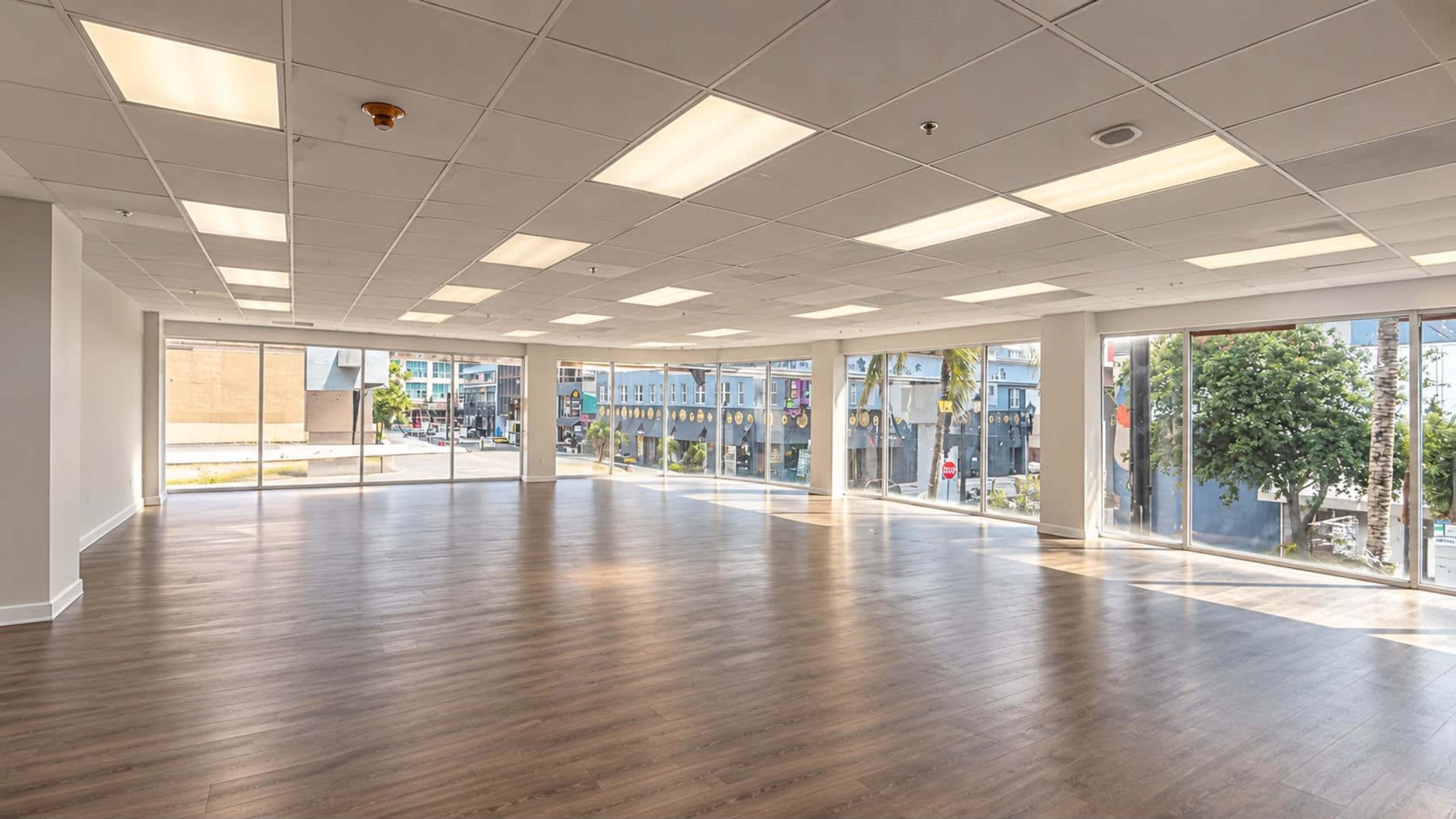 An empty commercial space features large windows allowing natural light, with a wooden floor and overhead fluorescent lighting.