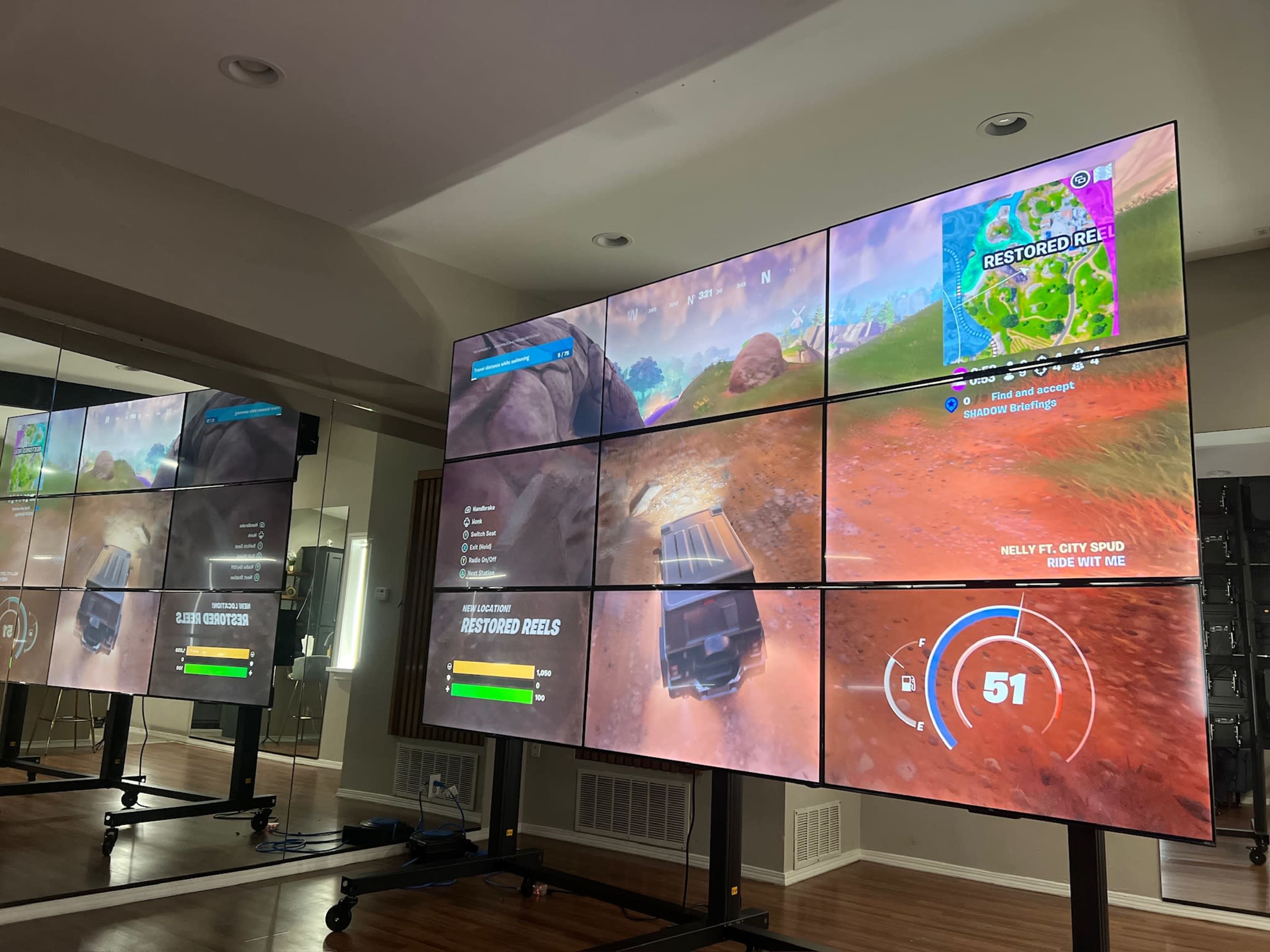 A large multi-screen setup displays a gameplay scene from a video game, showing various game stats and a landscape on each screen.